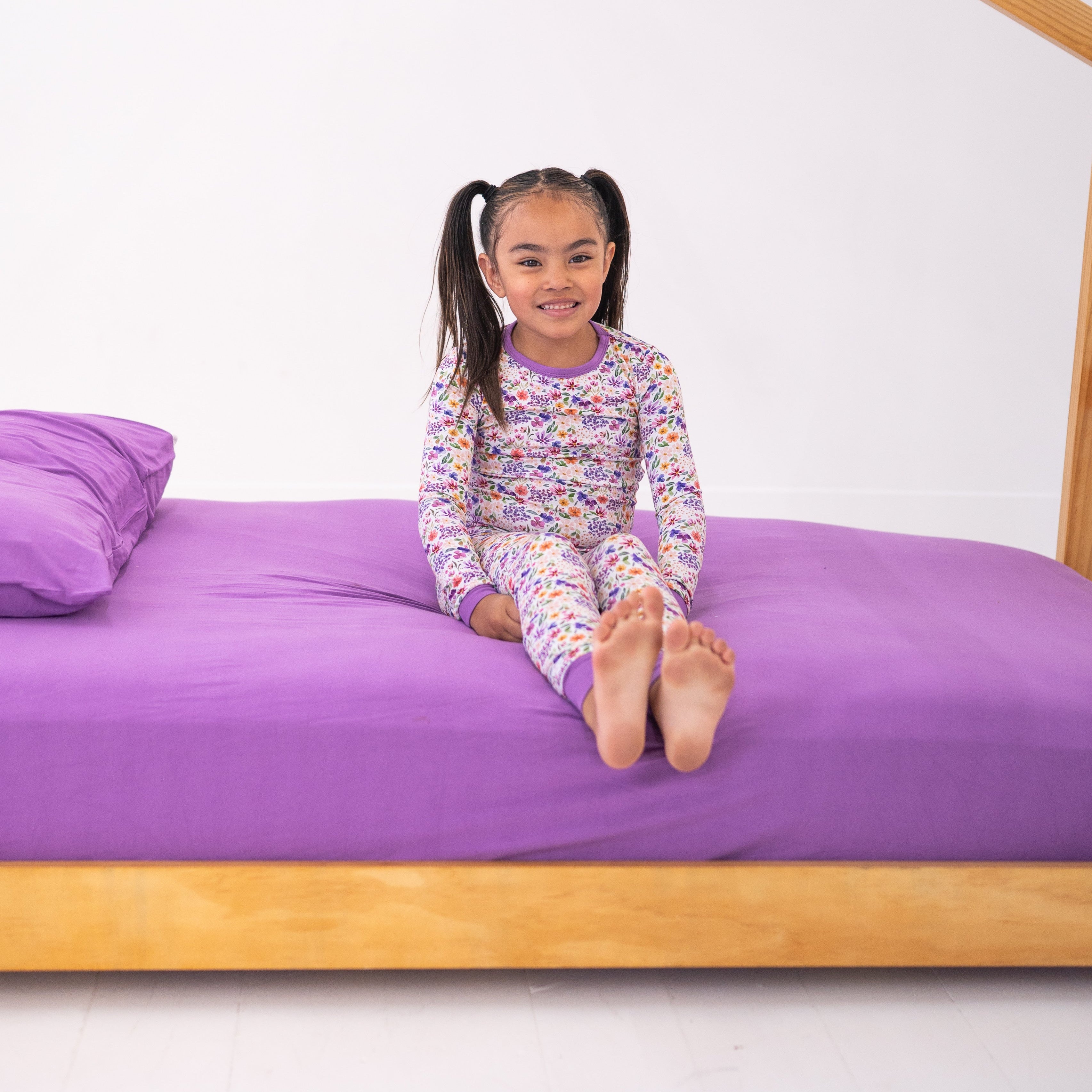 A young girl in floral pajamas sits on a purple twin bed with a pillow.