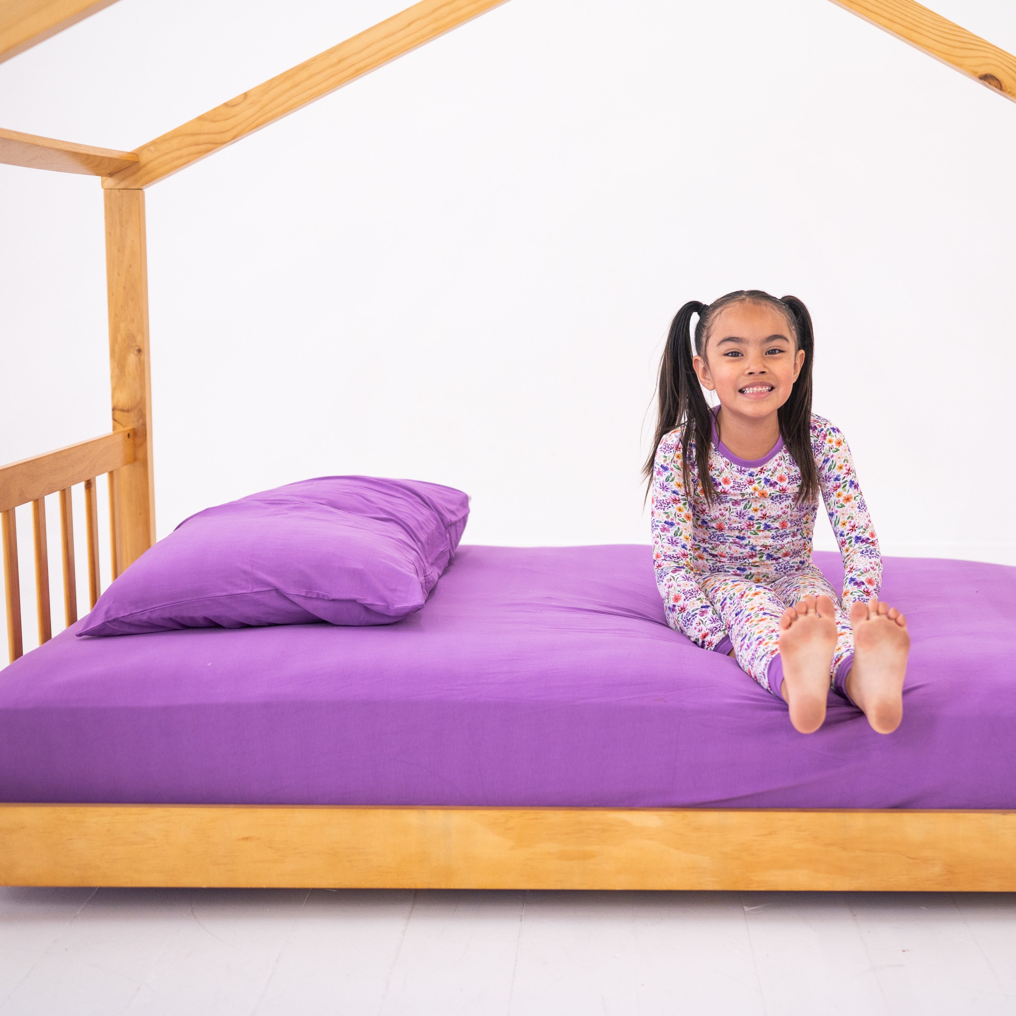 A young girl in floral pajamas sits on a twin bed with lavender sheets and a pillow.