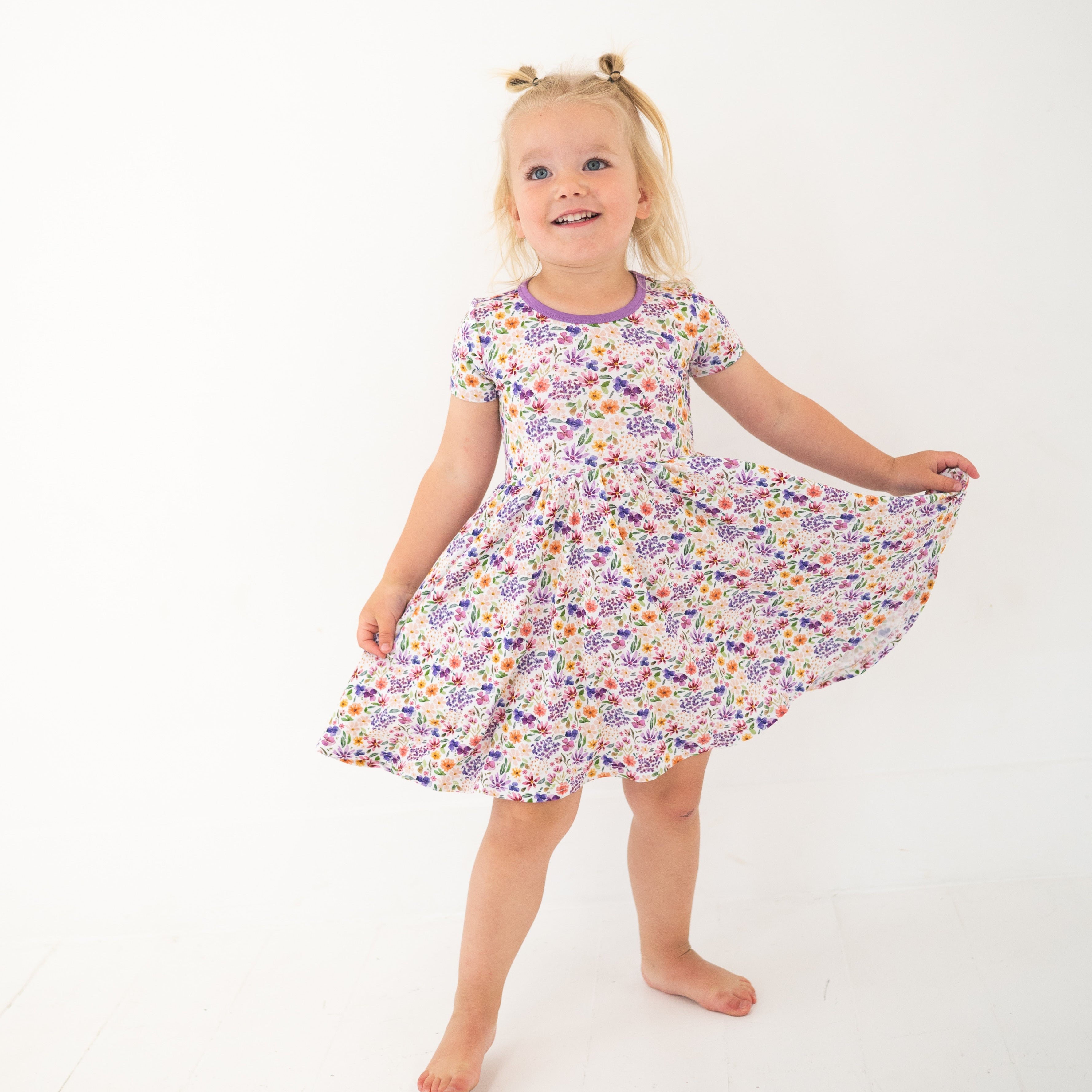A young girl in a white floral dress with purple trim twirls against a white background.
