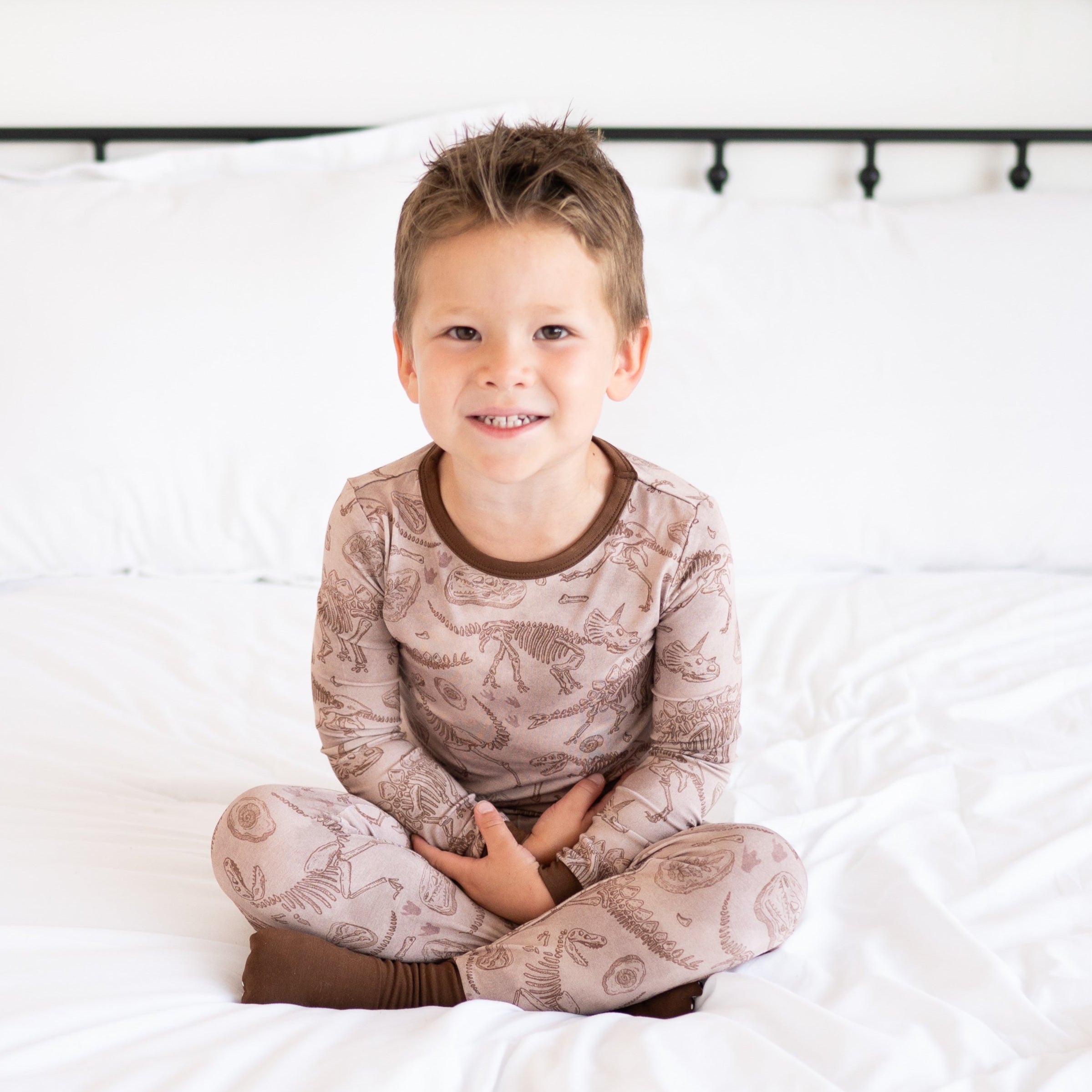 A young boy sits cross-legged on a white bed wearing a brown pajama set with a fossil print.
