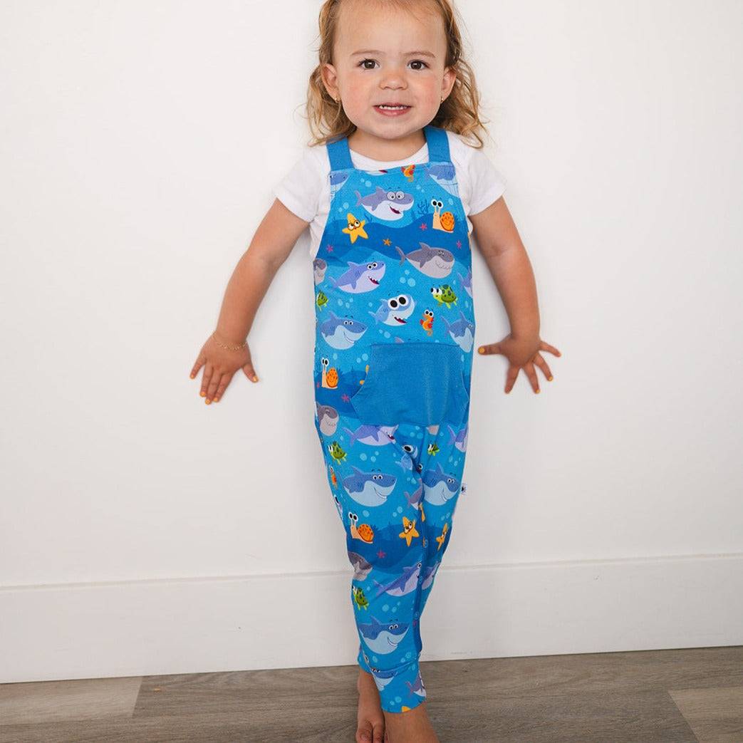 A toddler girl wears blue overalls with a Baby Shark print.