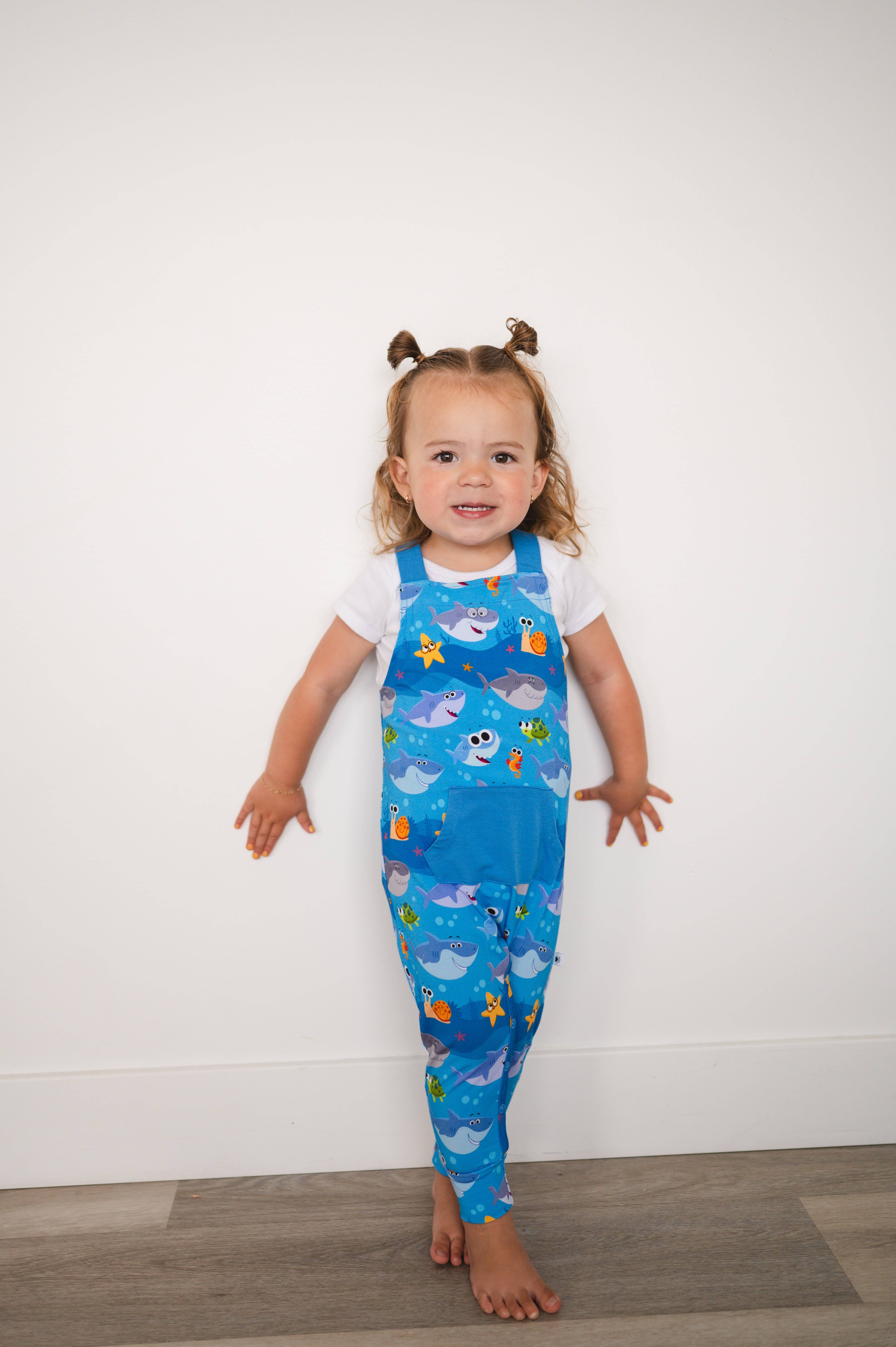 A toddler girl wears blue overalls with a Baby Shark print.