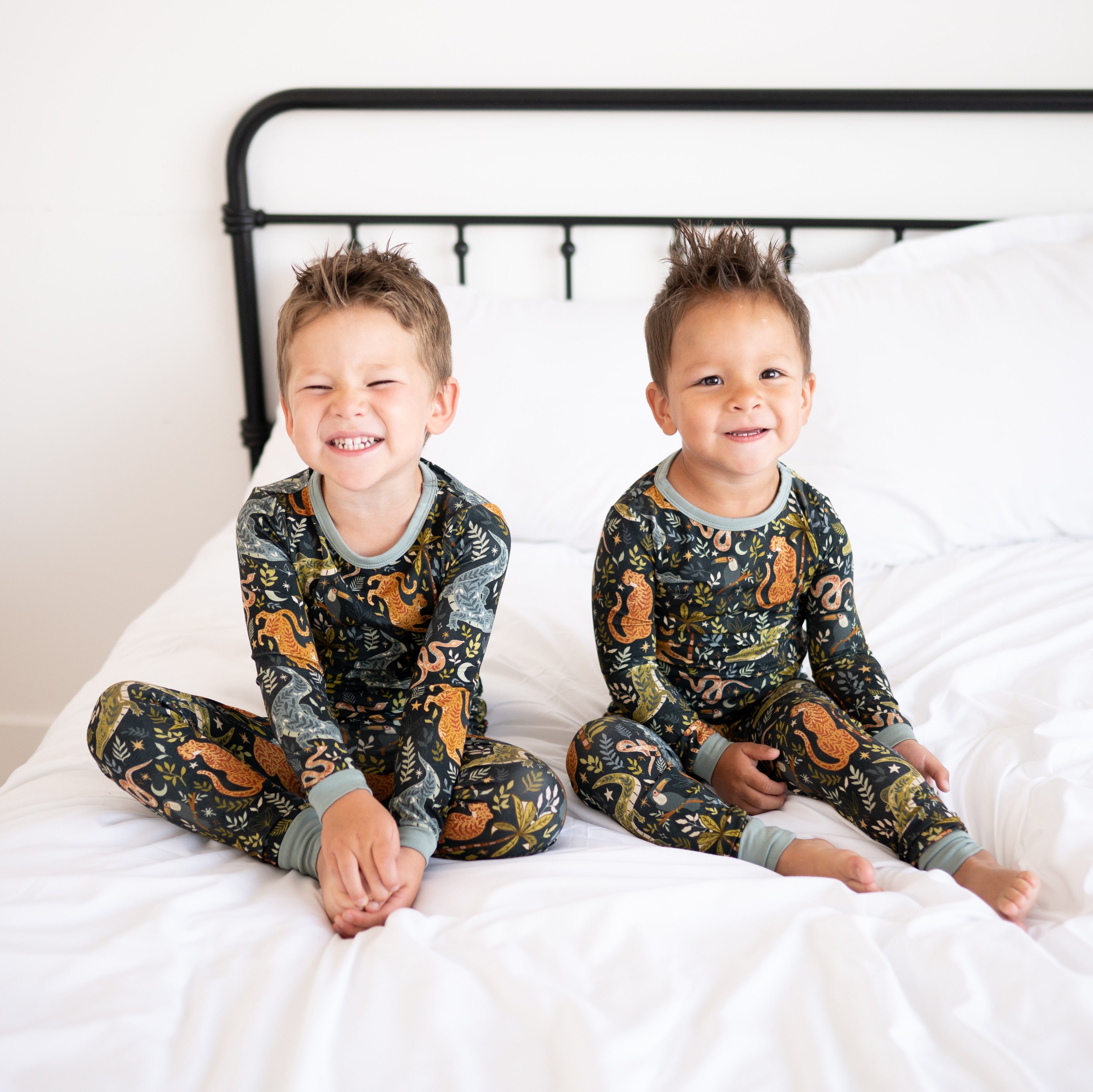 Two young boys in matching Night Jungle bamboo pajamas sit on a white bed.