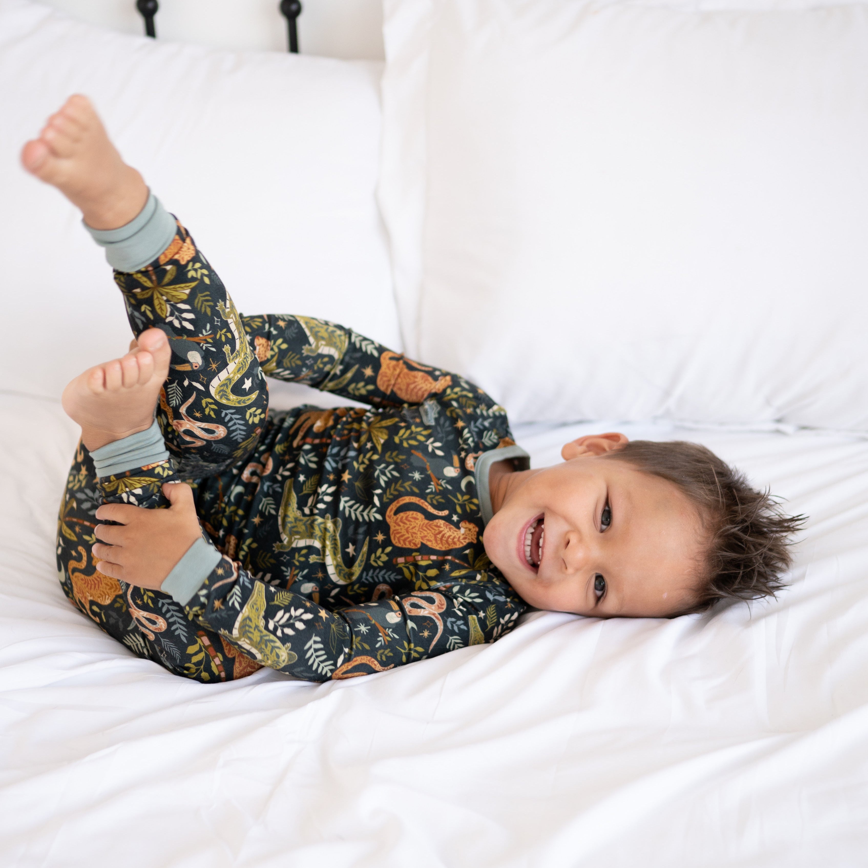 A young boy in a dark green pajama set with a jungle print smiles while lying on a white bed.
