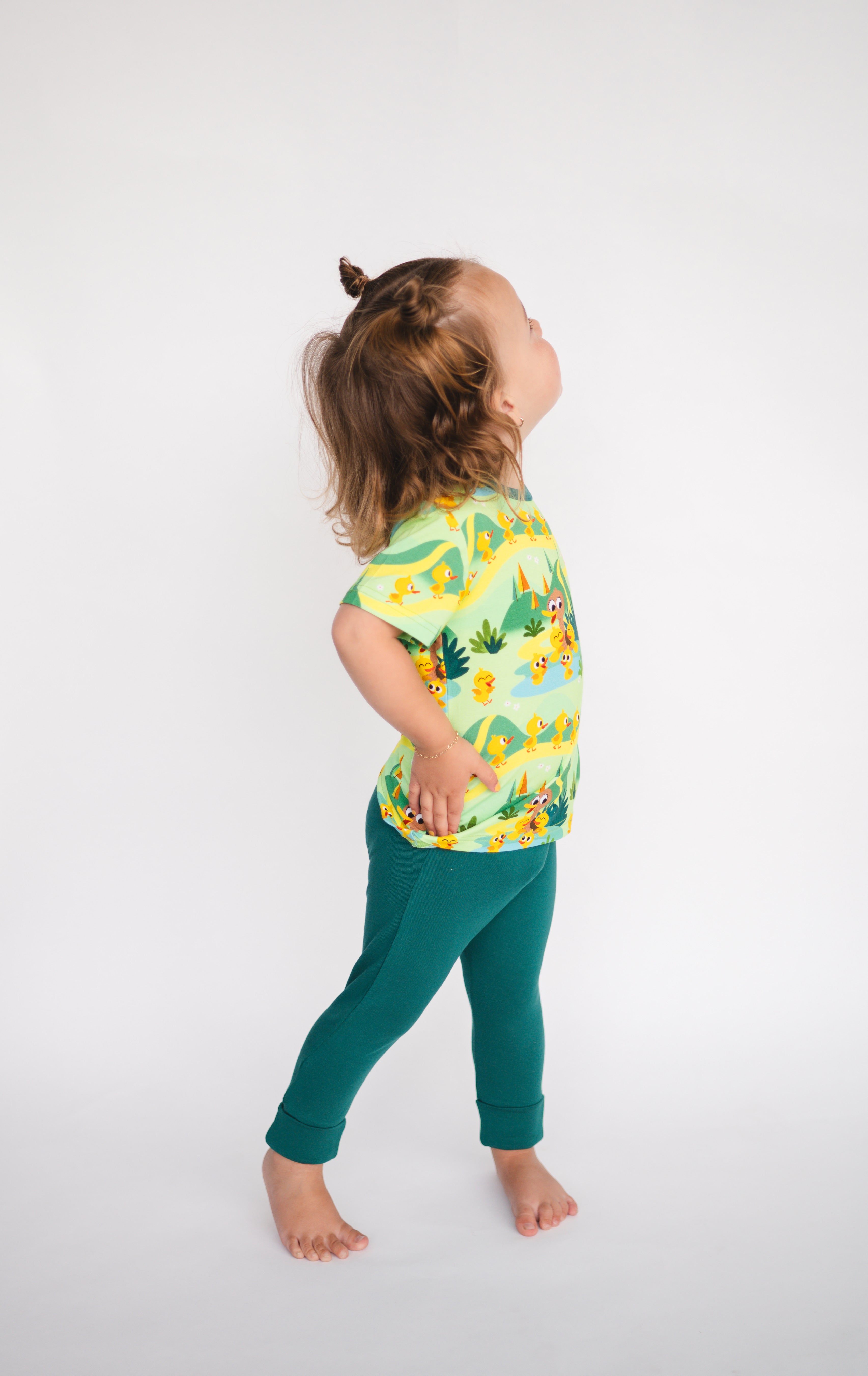 A toddler wears green pants and a green duck-themed shirt.