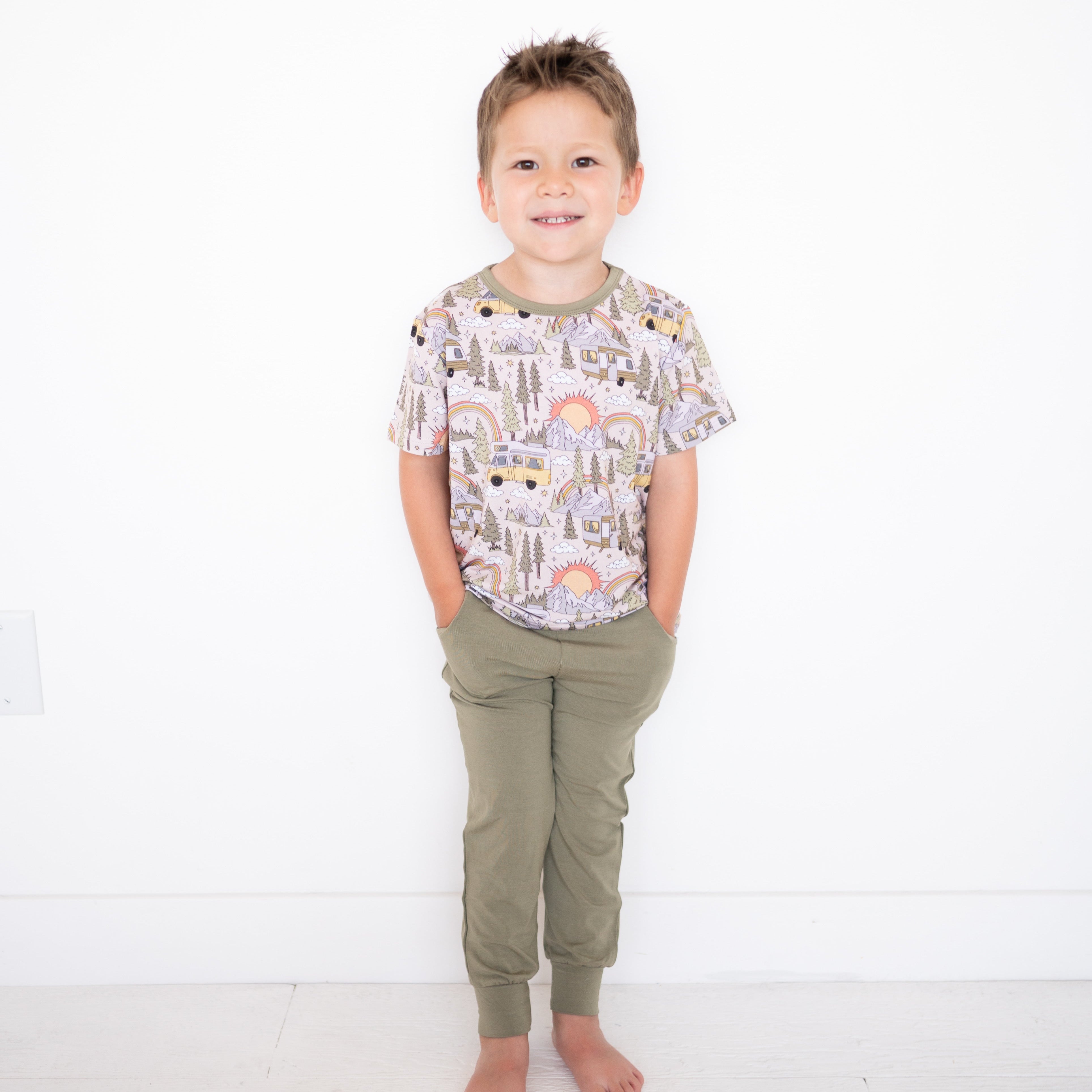 A young boy wears a light pink t-shirt with a vintage camper and mountain print and olive green joggers.