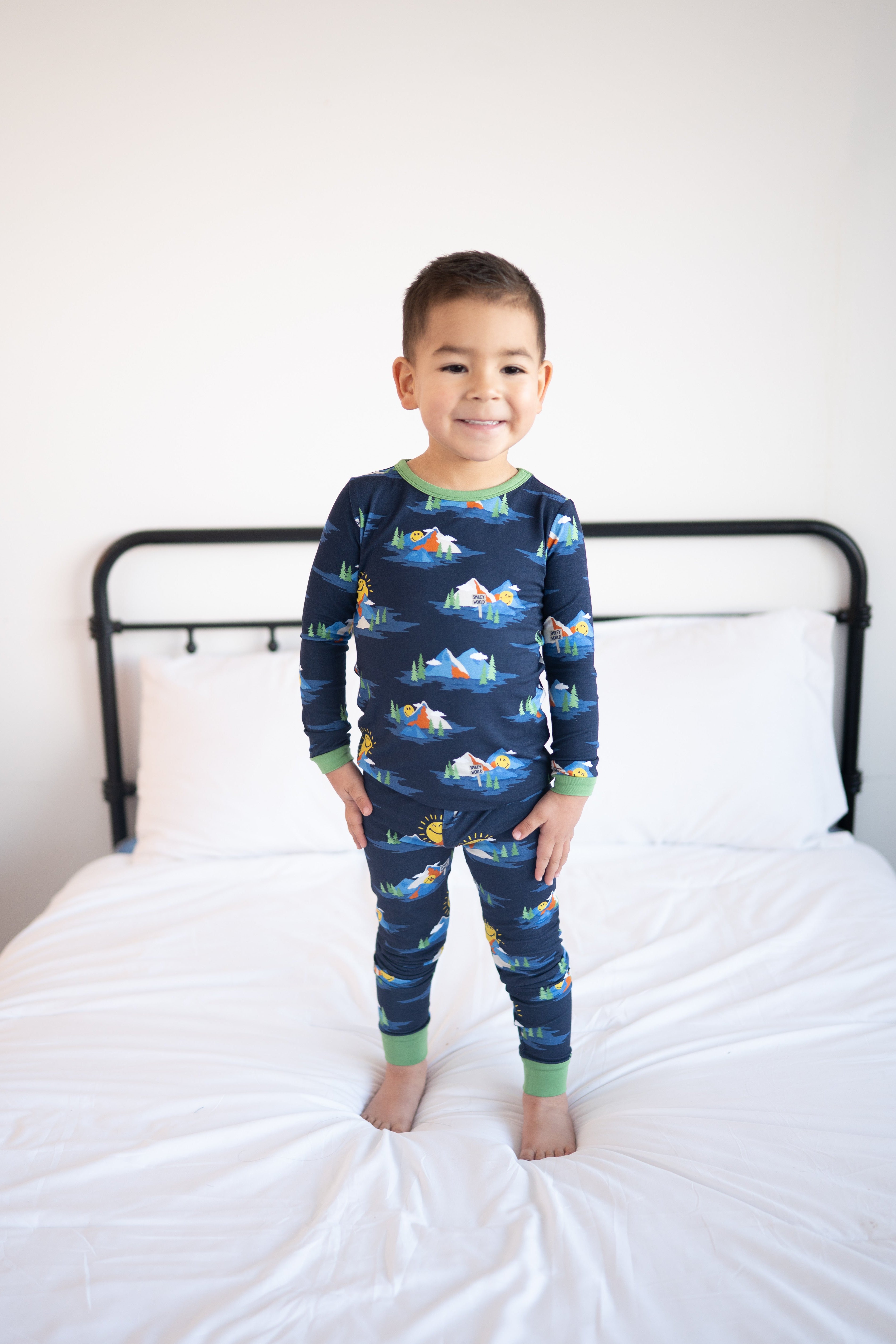 A young boy stands on a bed wearing navy blue pajamas with a Mount SmileyWorld print.