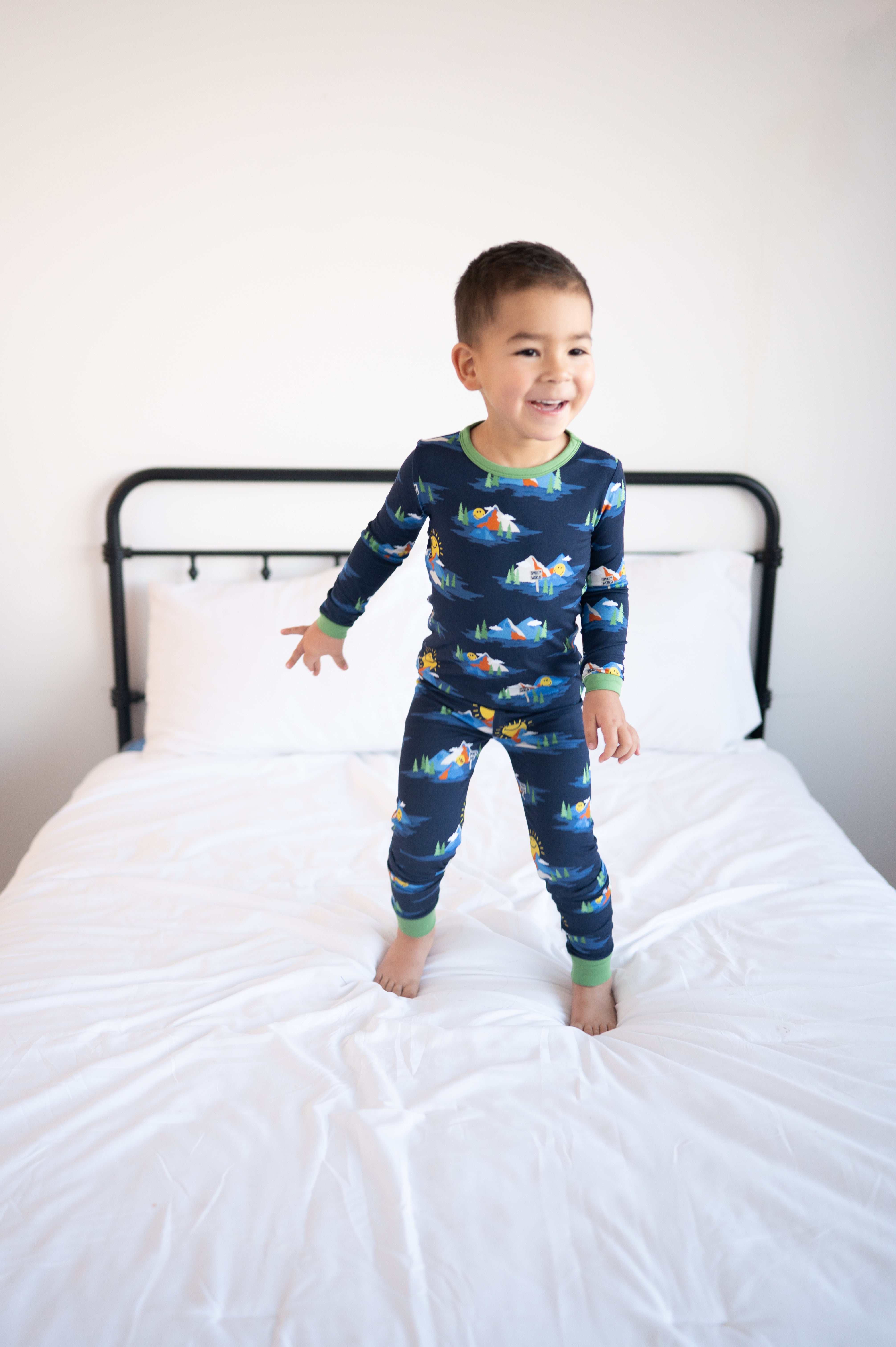 A young boy in navy blue pajamas with a SmileyWorld mountain print stands on a white bed.