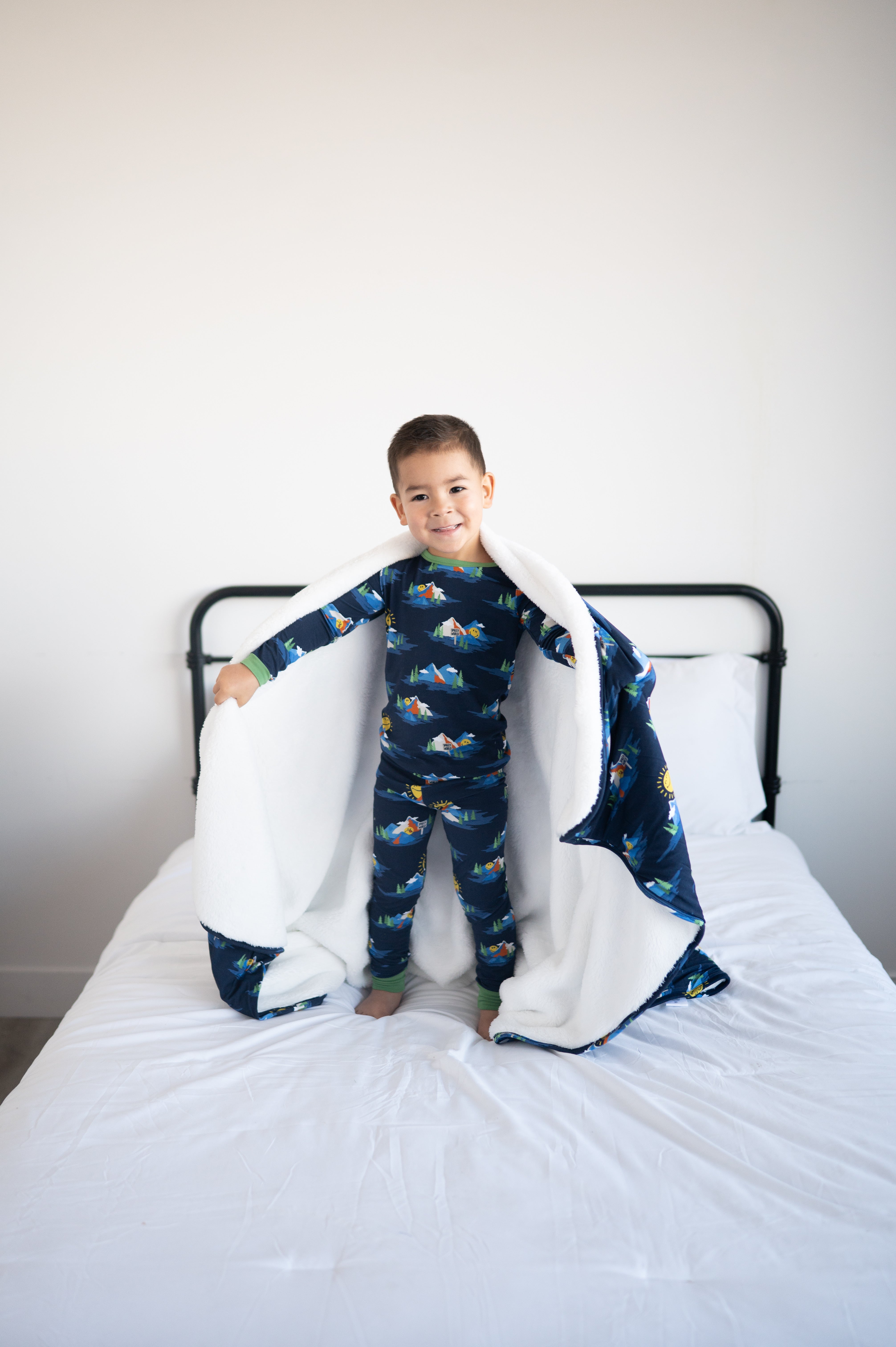 A young boy in SmileyWorld pajamas and a fluffy blanket stands on a bed.
