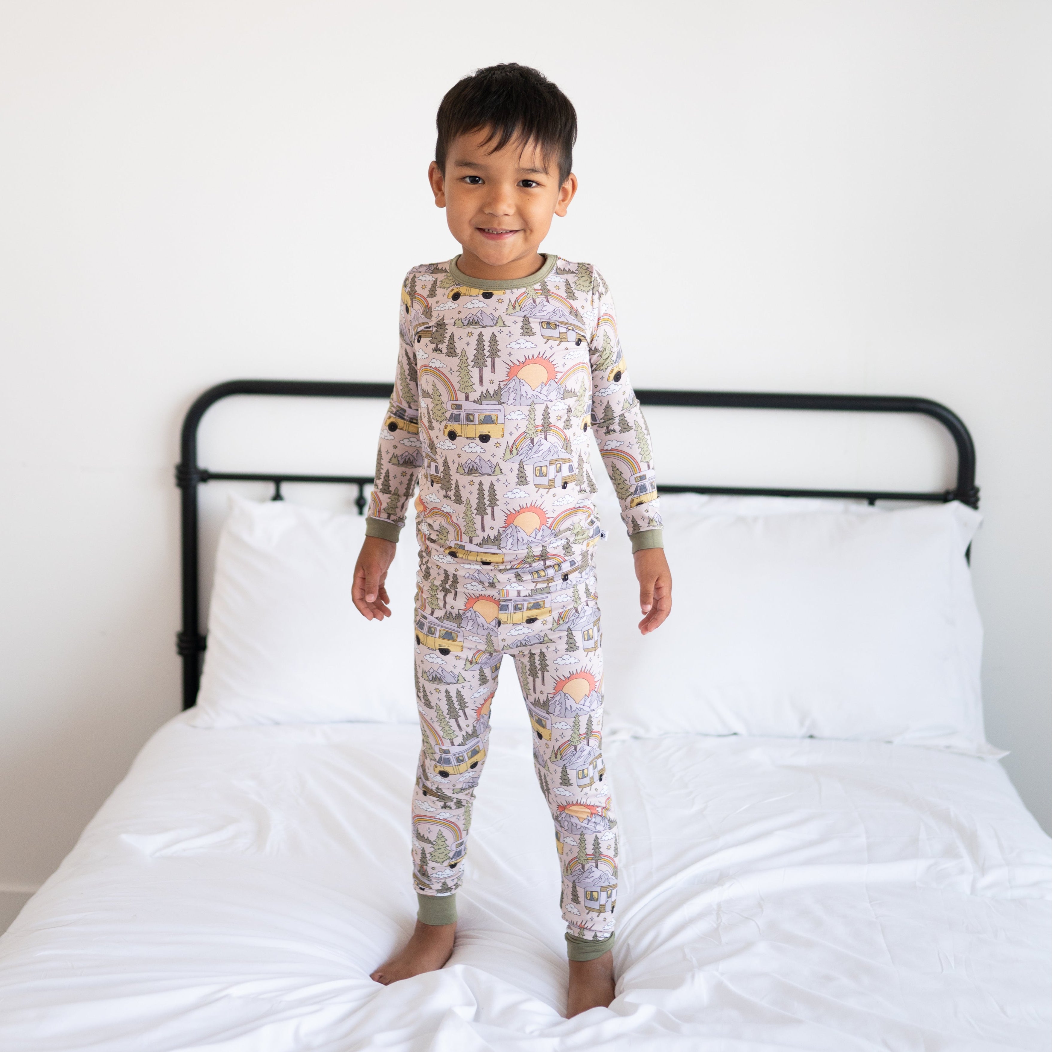 A young boy stands on a bed wearing a long-sleeved pajama set with a vintage camper and nature print.
