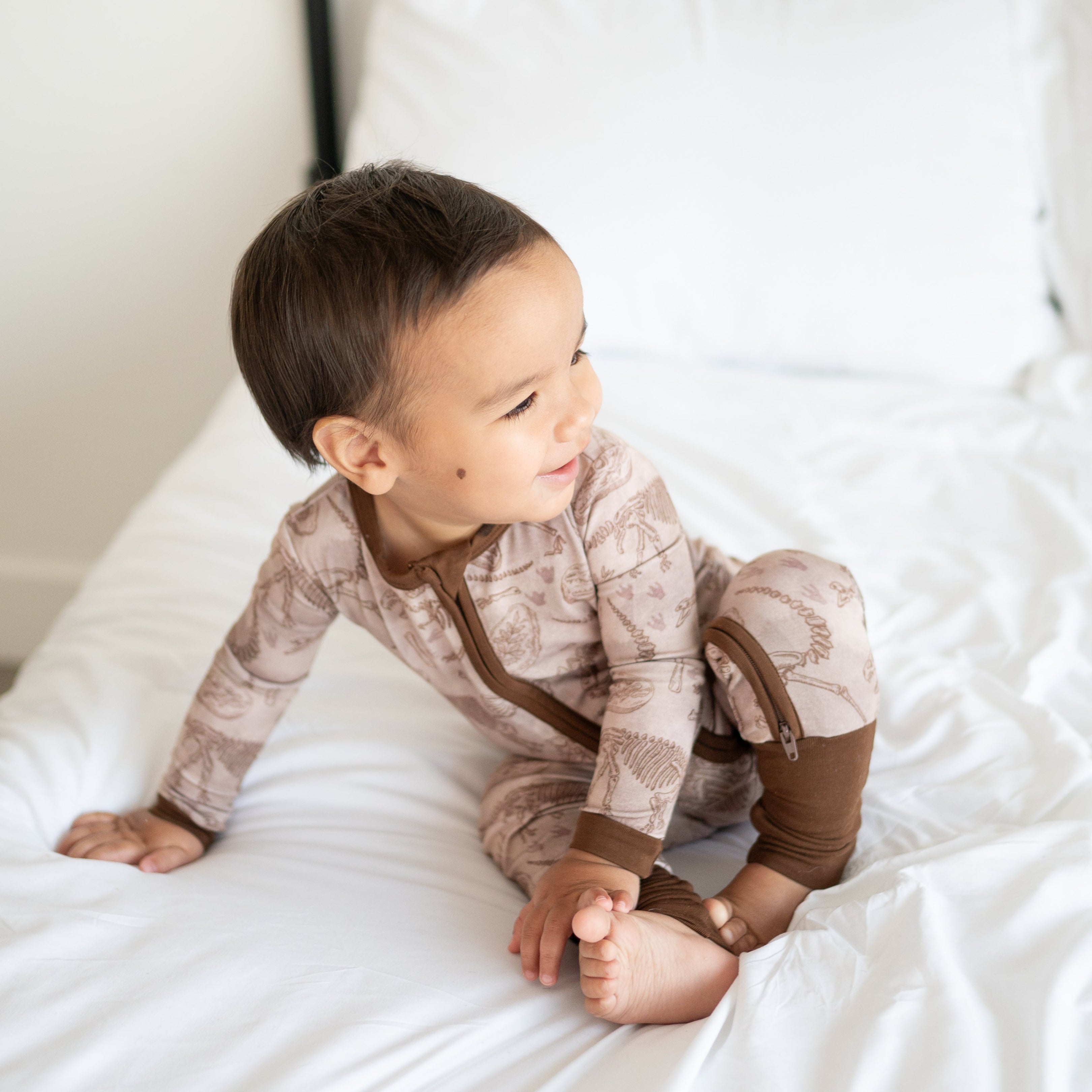 A baby wearing a tan bamboo pajama set with a fossil print sits on a white bed.