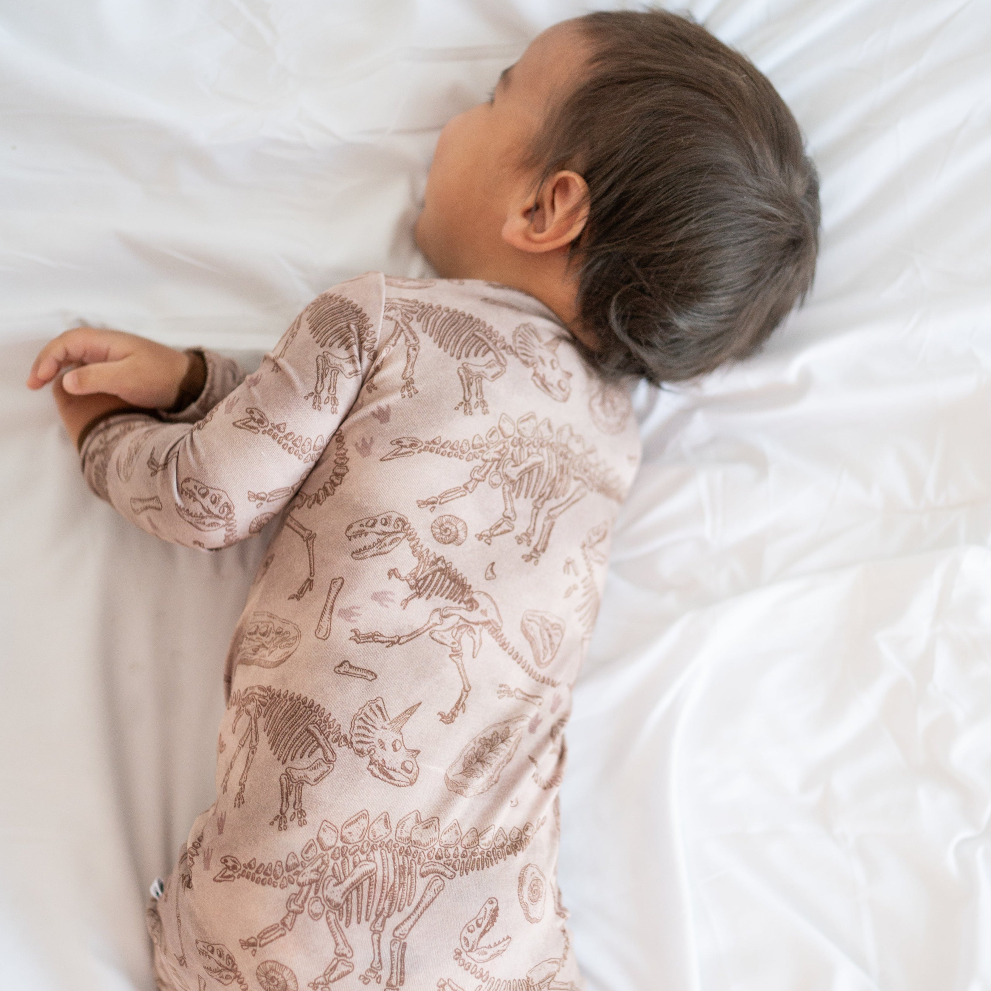 A baby sleeps on a white sheet wearing a tan pajama set with a fossil print.