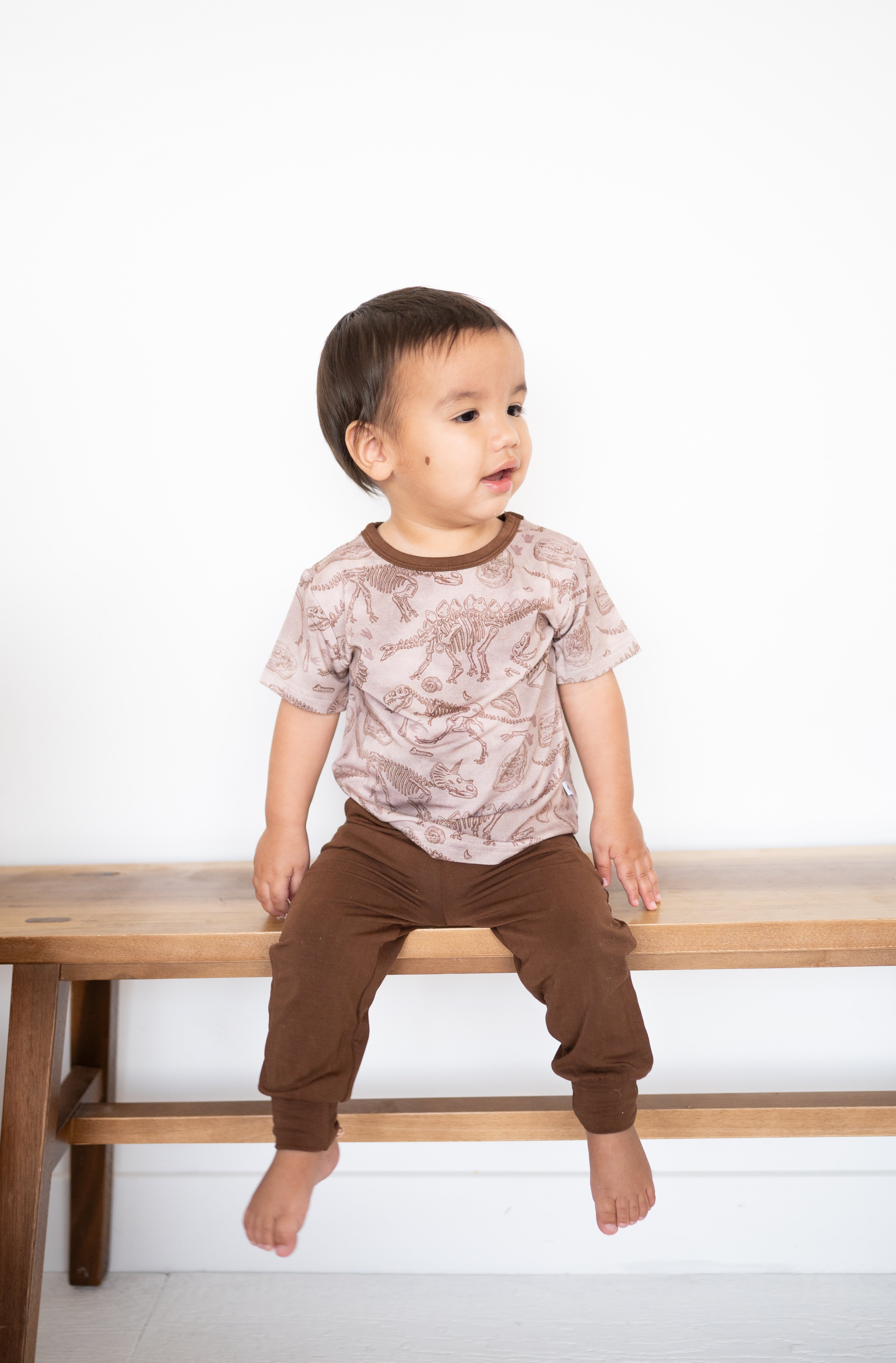 A toddler boy in a dinosaur print shirt and brown joggers sits on a wooden bench.