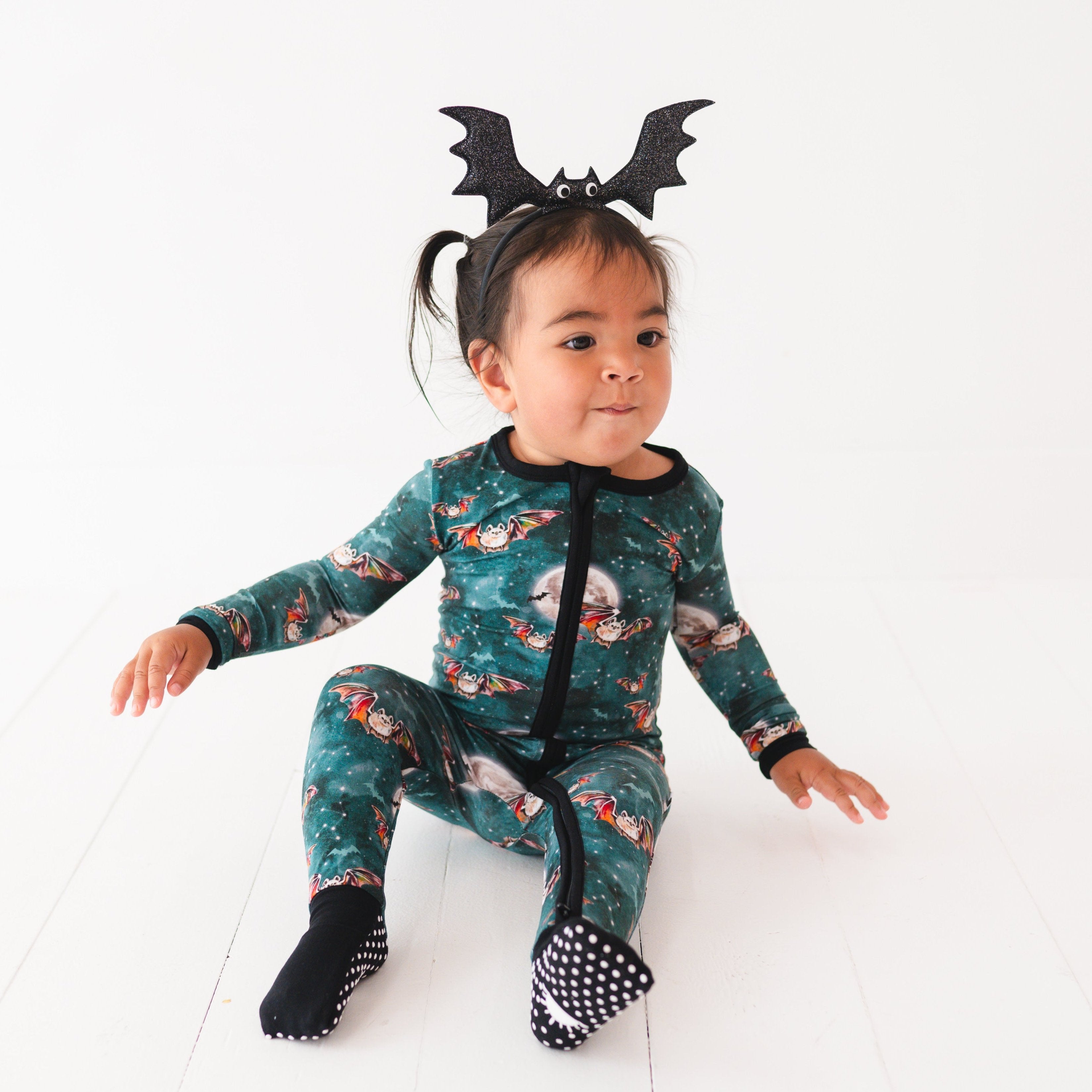 A baby wearing a teal pajama set with a bat print and a bat headband.