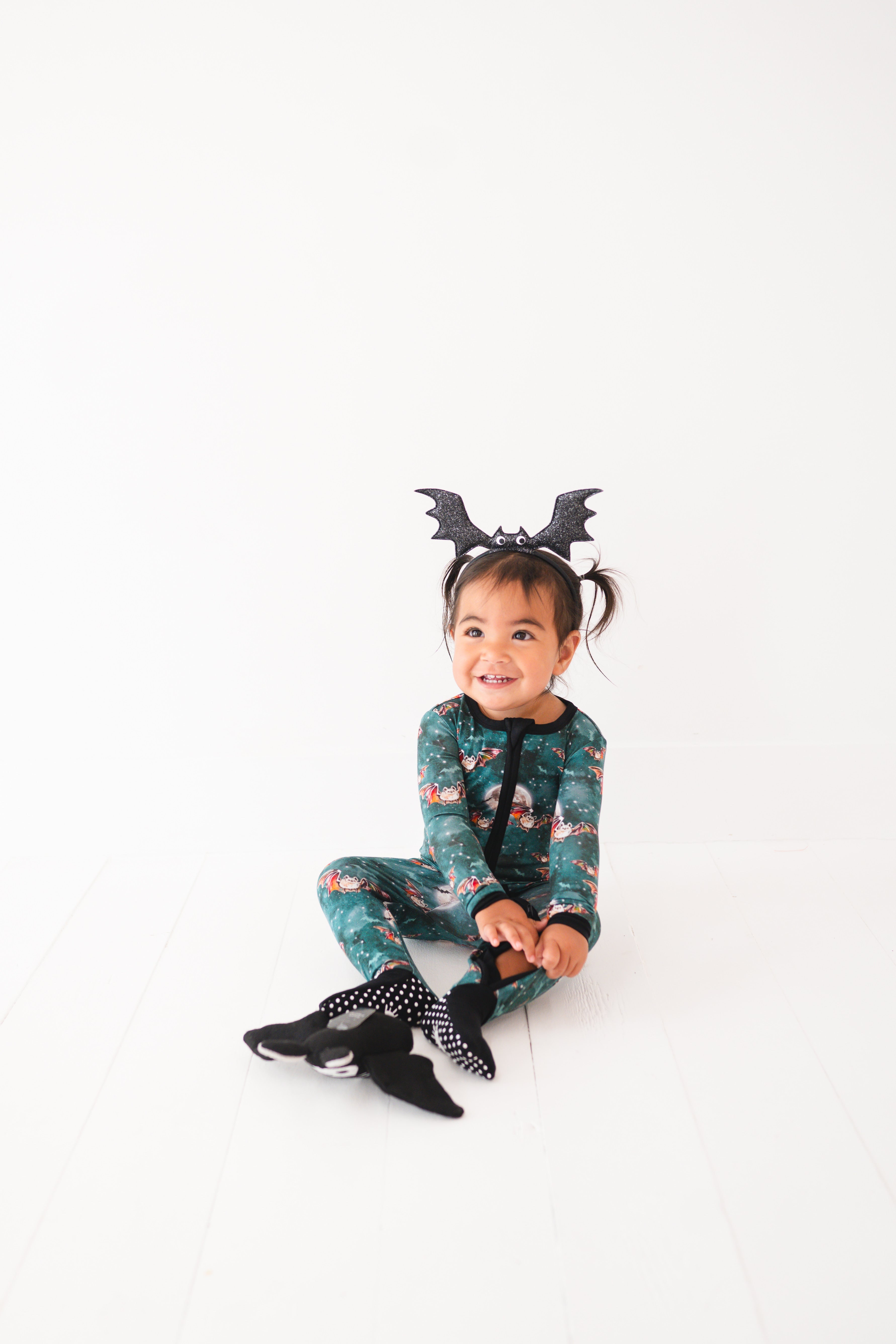 A toddler girl wears a green bamboo pajama set with bats and moons, and a bat headband.