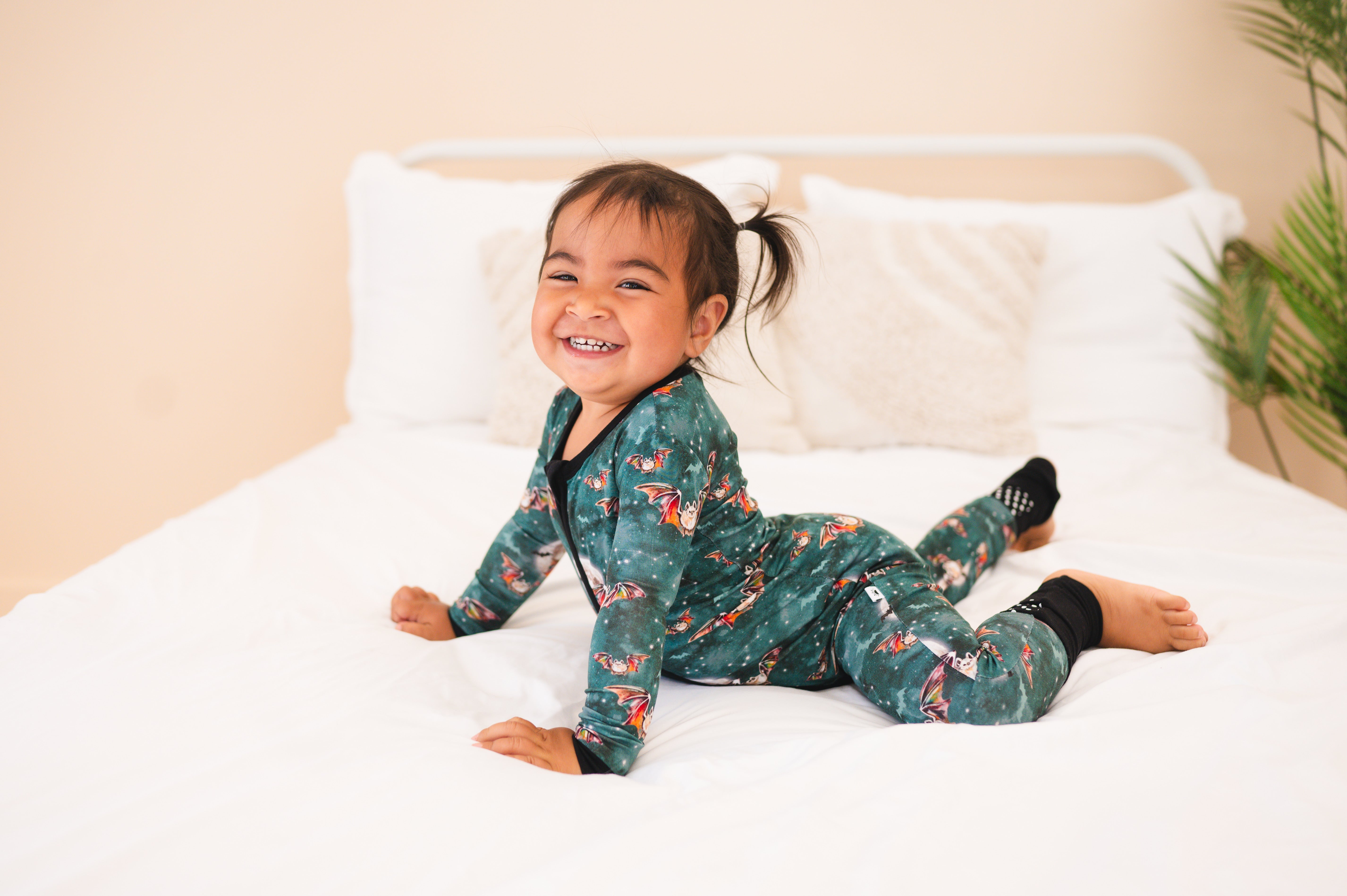 A baby in green pajamas with a bat print smiles on a white bed.