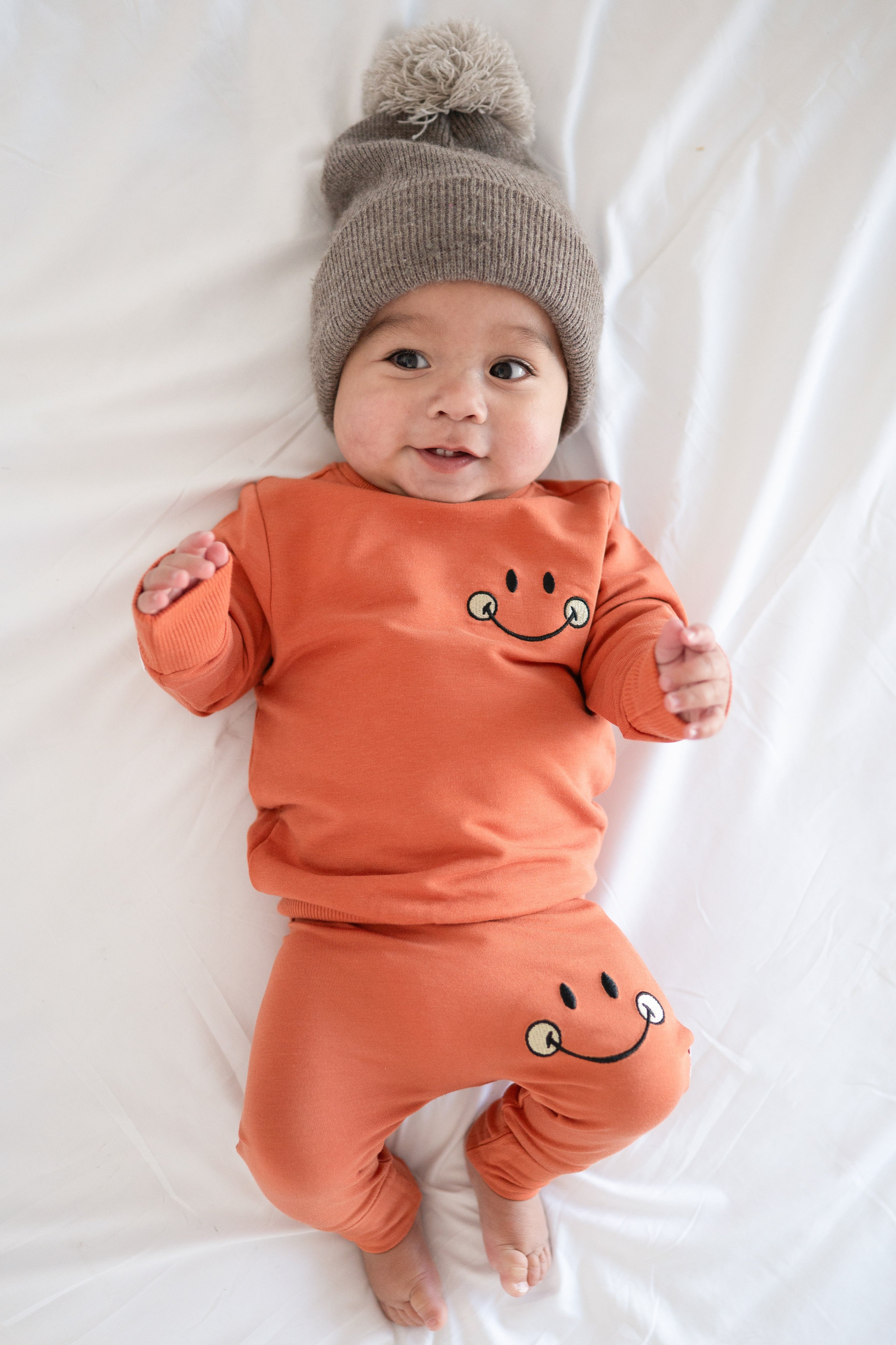 Baby in orange smiley face sweatshirt and pants with a knit hat.