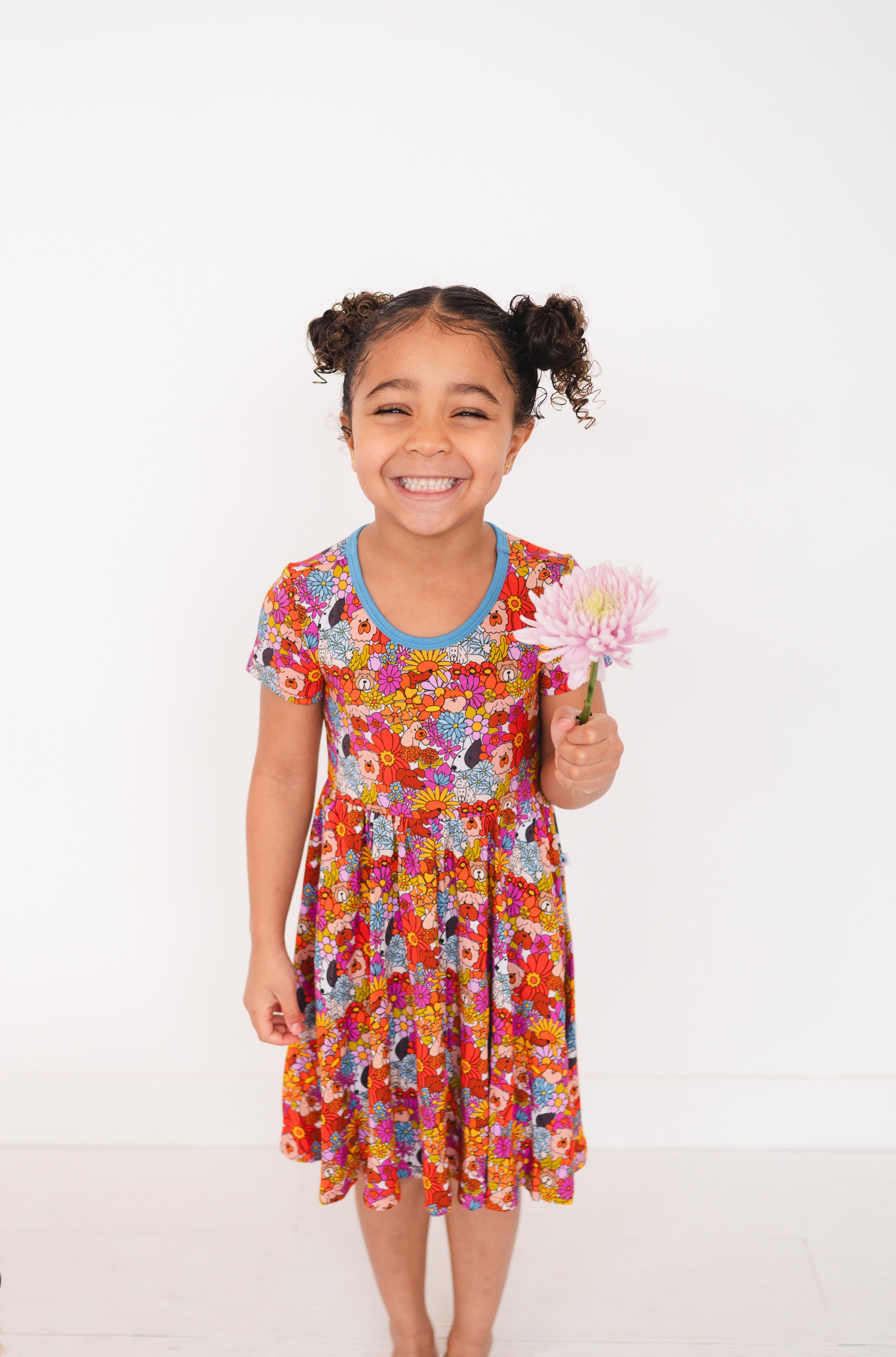 A young girl smiles holding a pink flower while wearing a colorful floral dress.