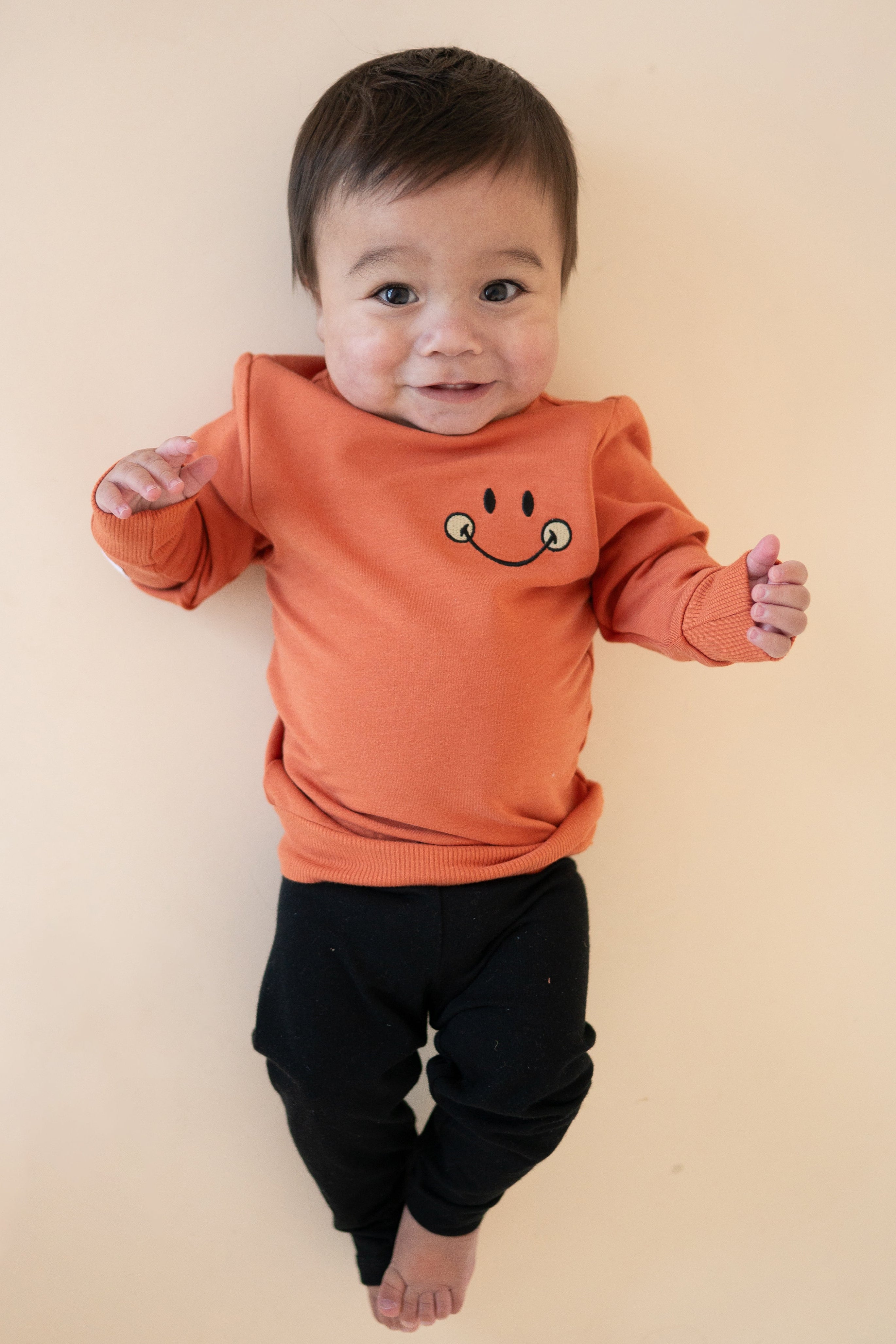 Baby in a rust-colored crewneck sweatshirt with a smiley face graphic.