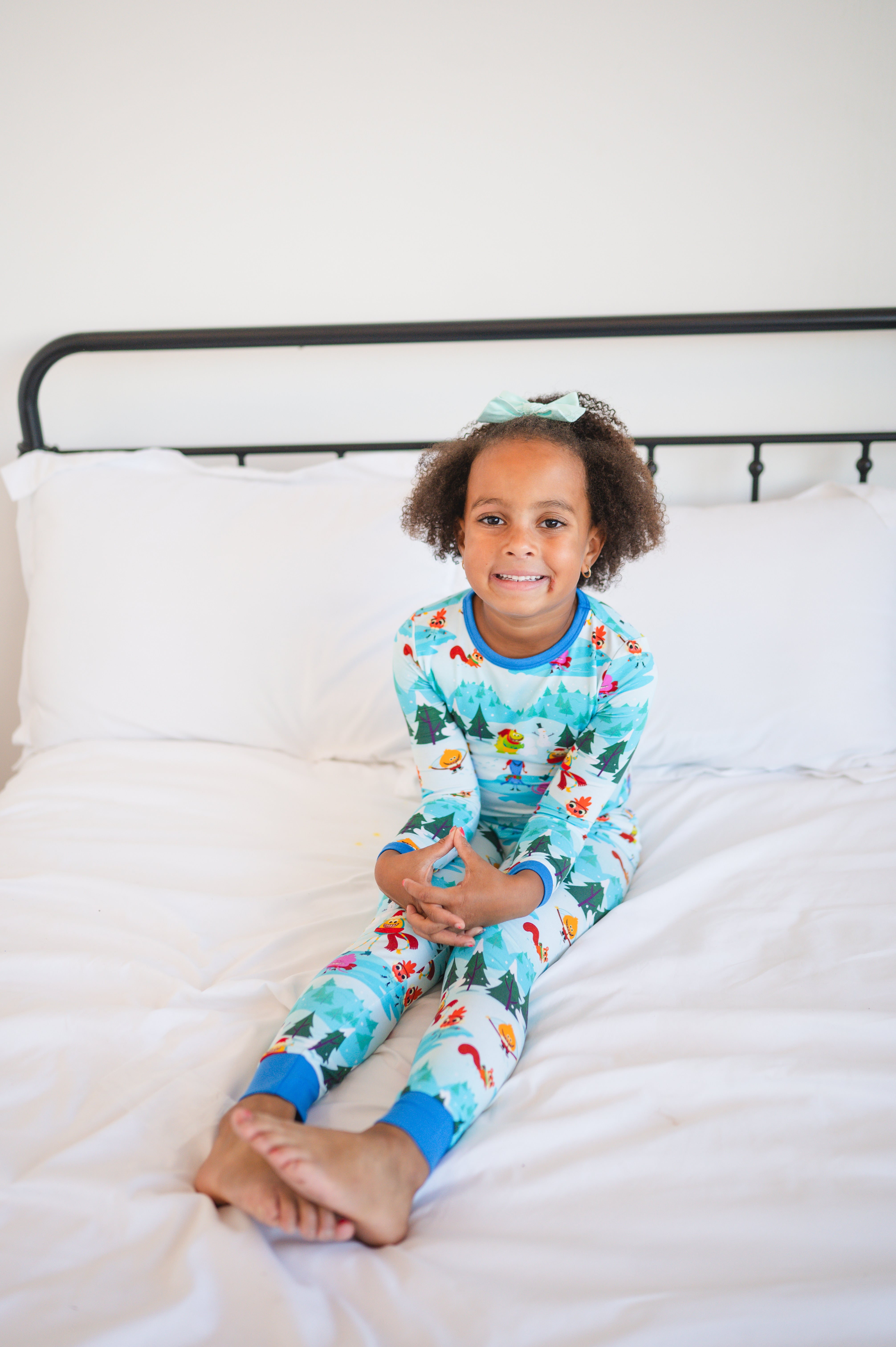 A young girl in blue pajamas with a winter forest print sits on a white bed.
