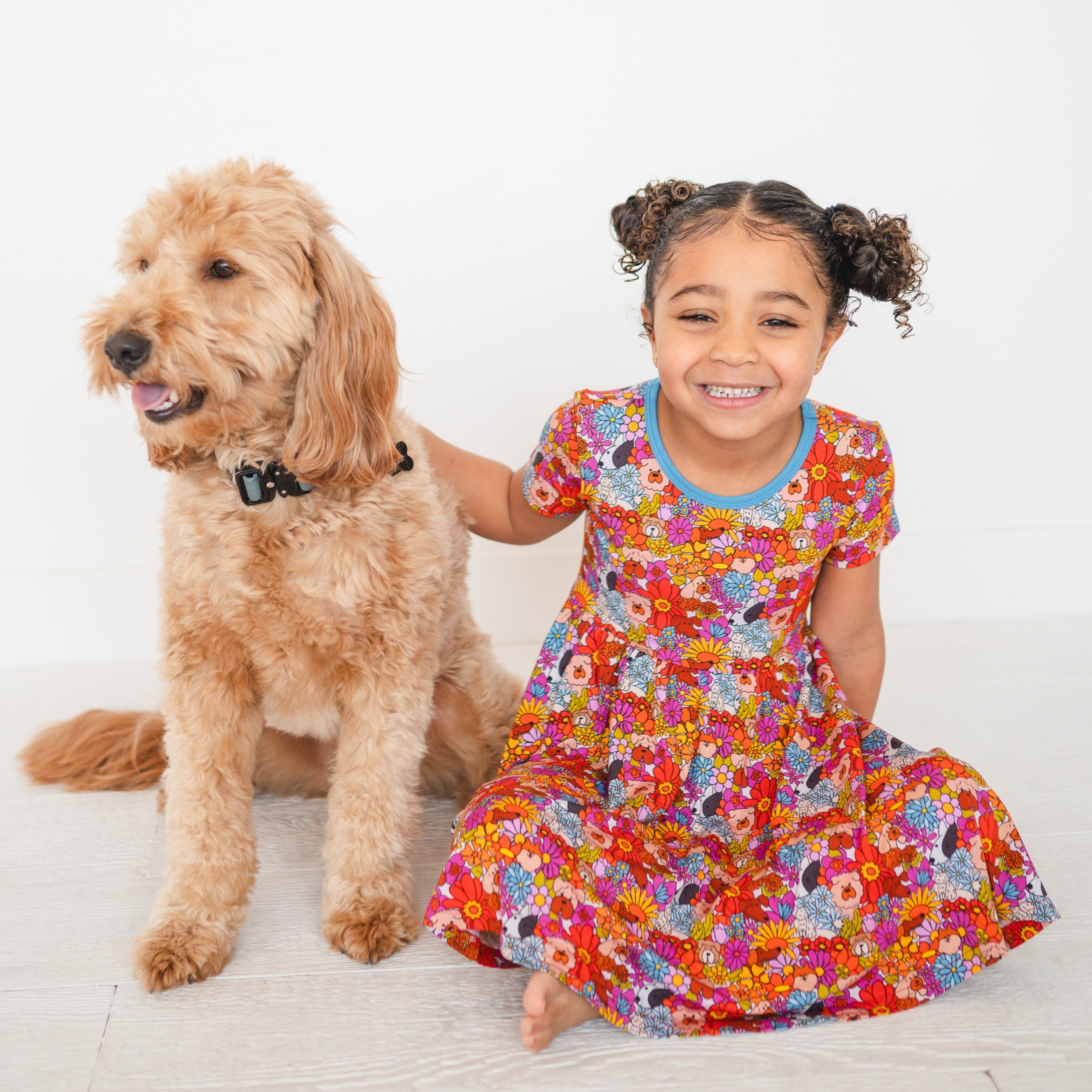 A young girl in a colorful floral dress sits next to a golden doodle dog.