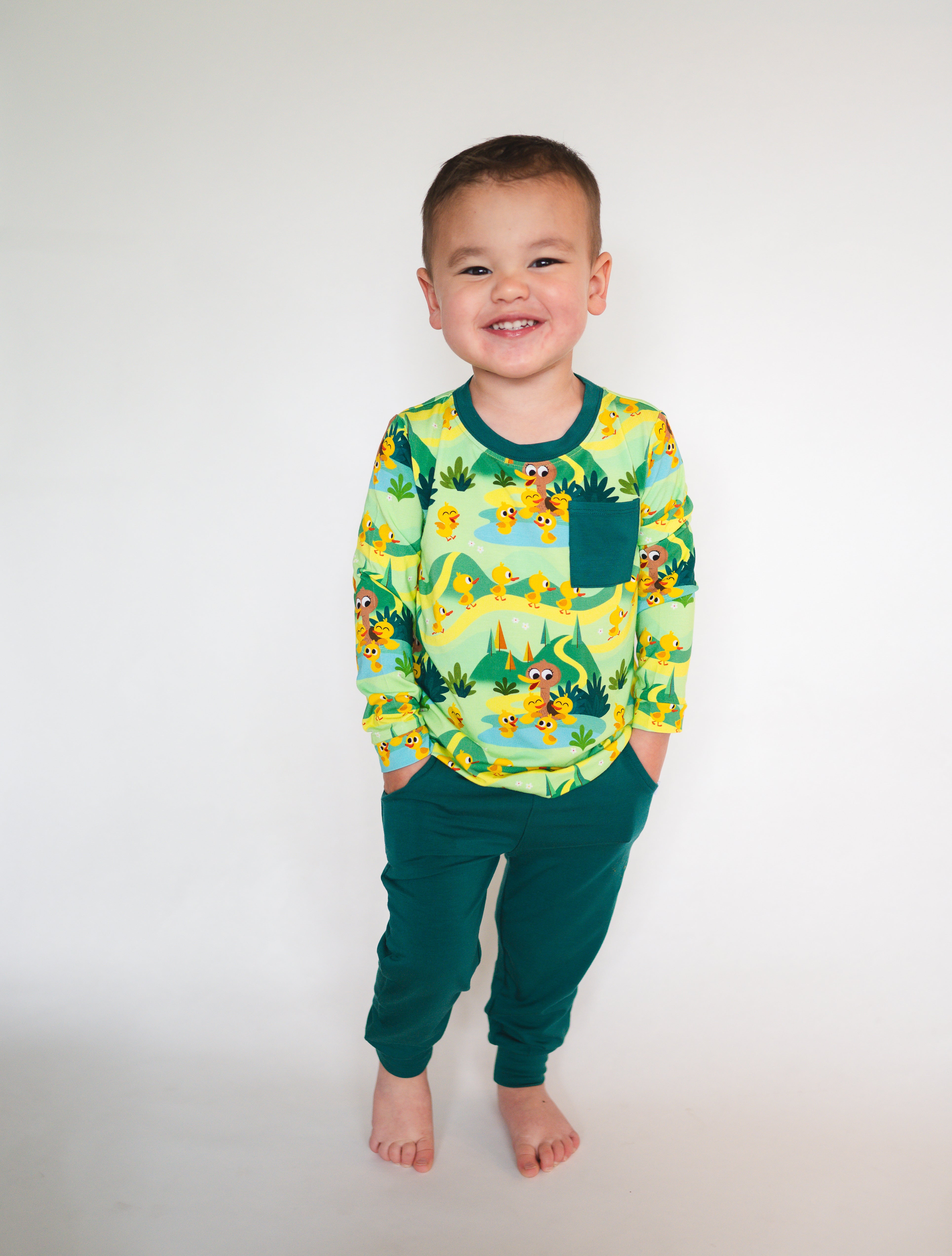 A young boy smiles wearing a green long-sleeved shirt with a duck pond print and green joggers.