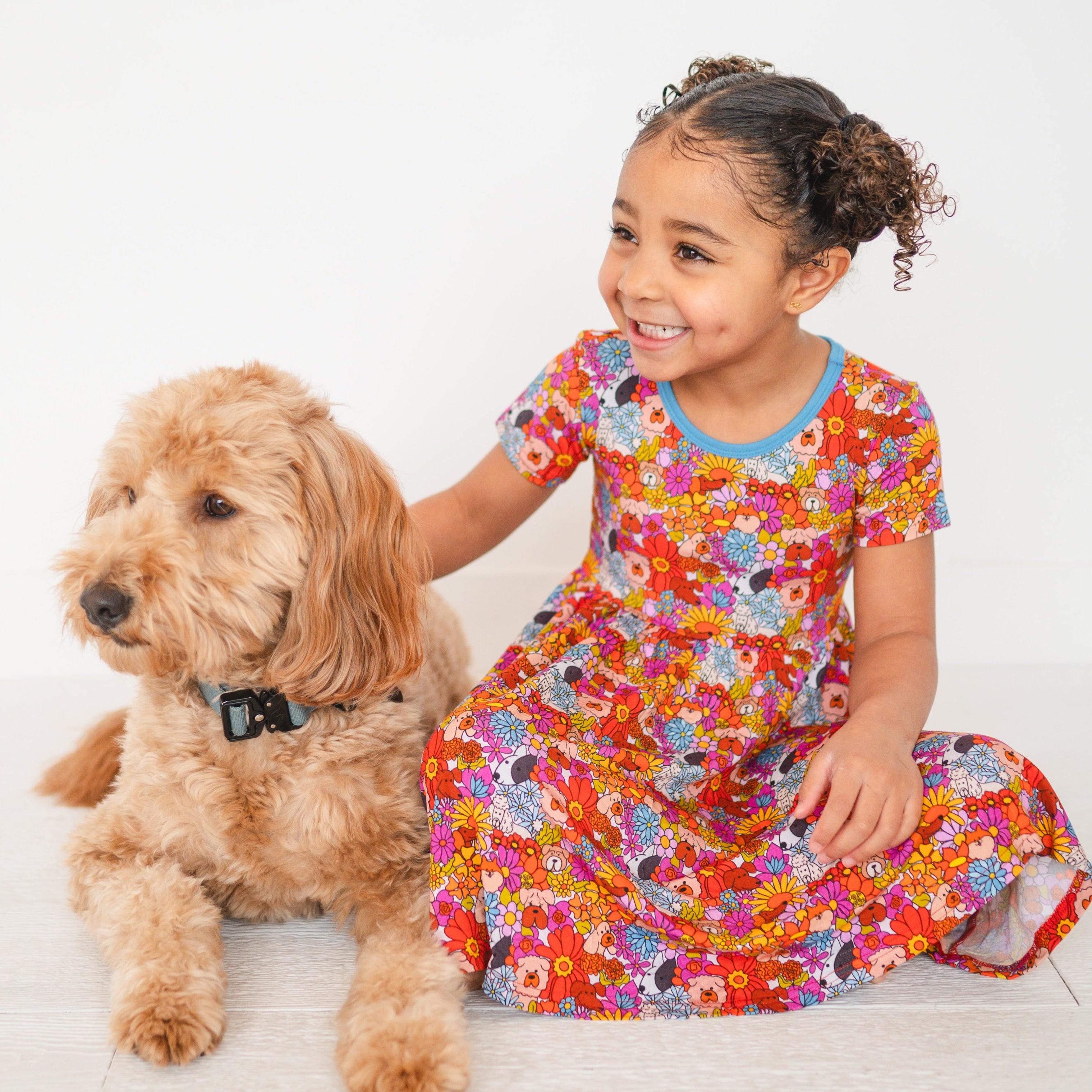 A young girl in a colorful floral and dog print dress sits next to a golden doodle dog.