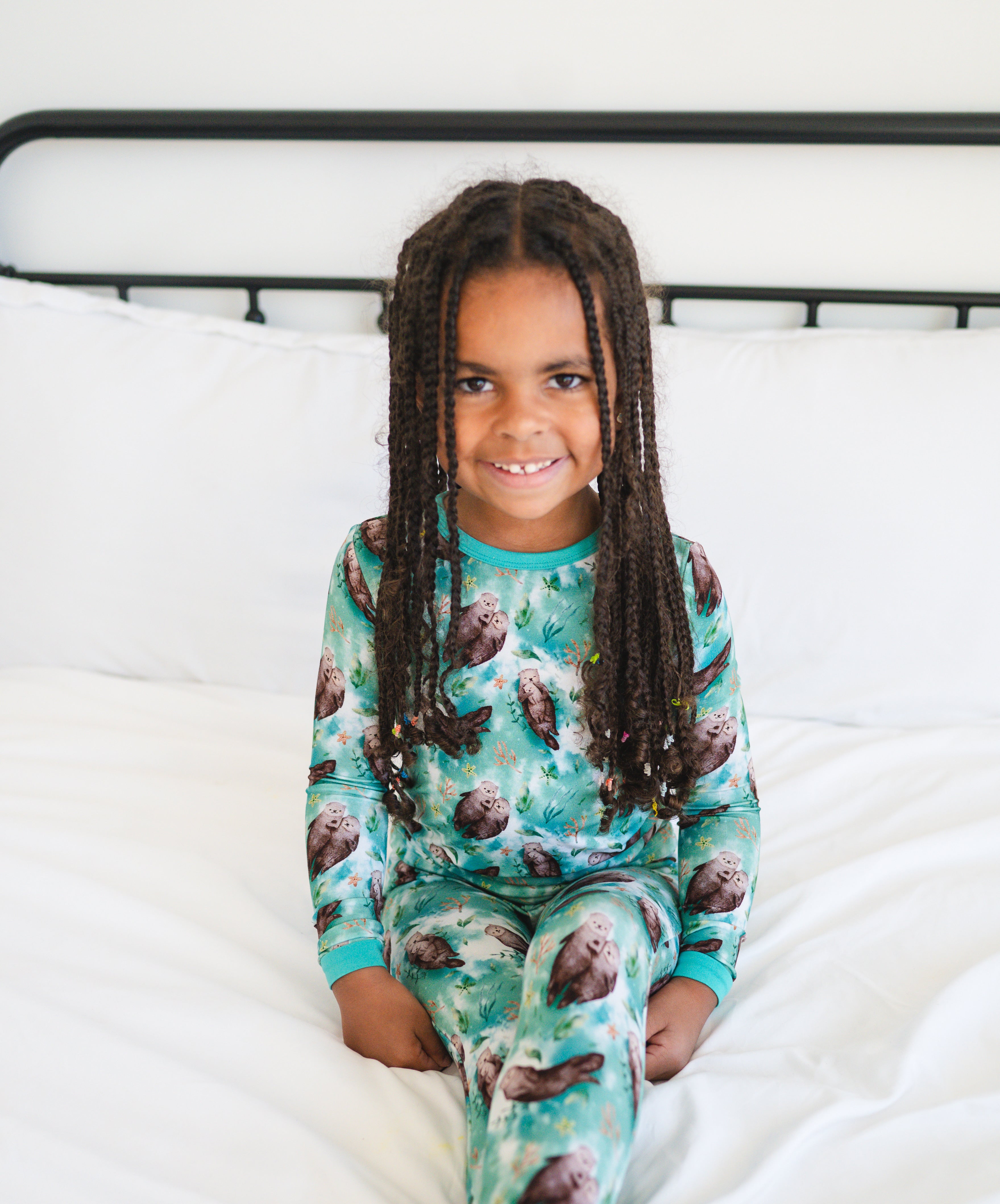 A young girl with braided hair smiles while wearing a teal pajama set with an otter print.