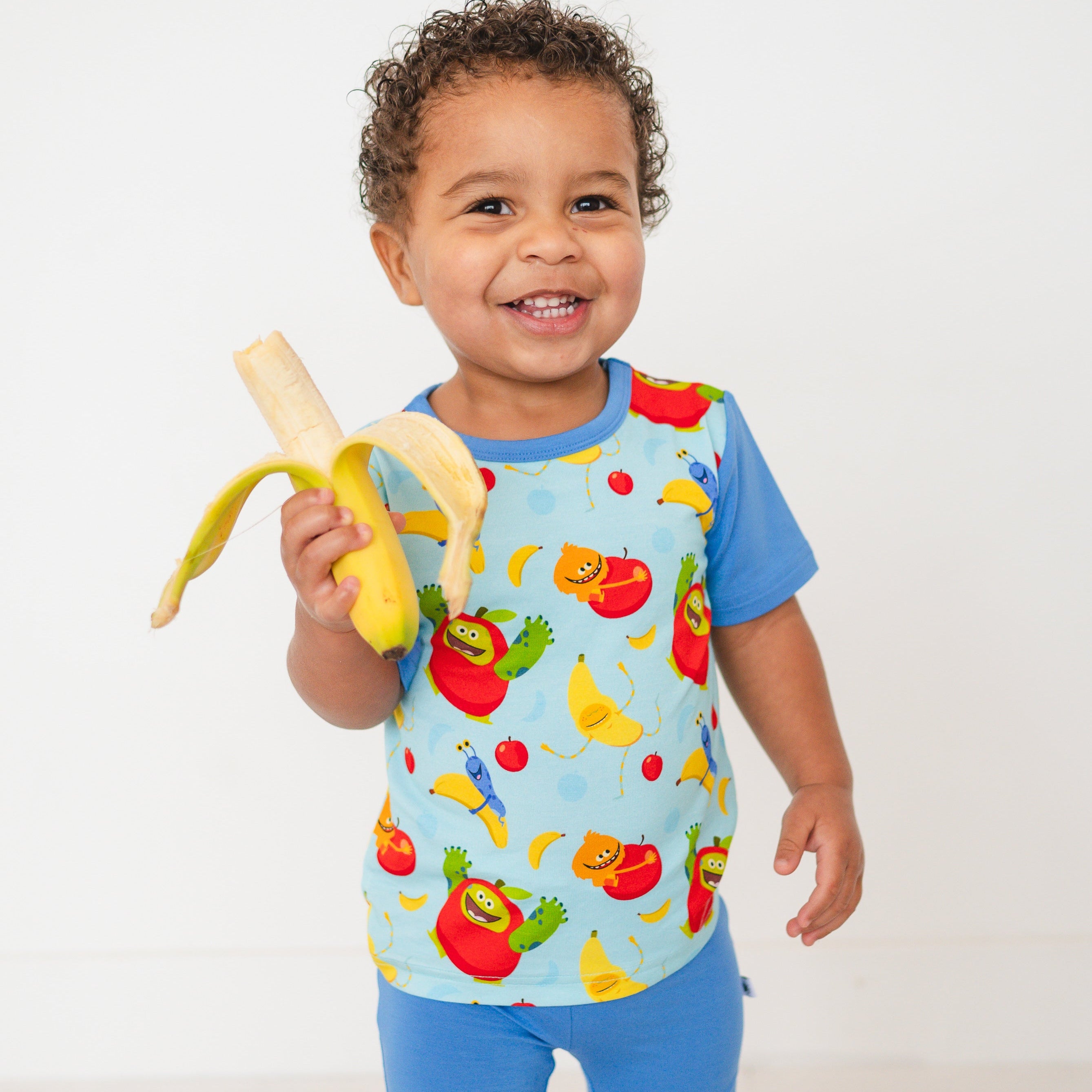 A smiling toddler holds a peeled banana while wearing a light blue t-shirt with cartoon apples and bananas.