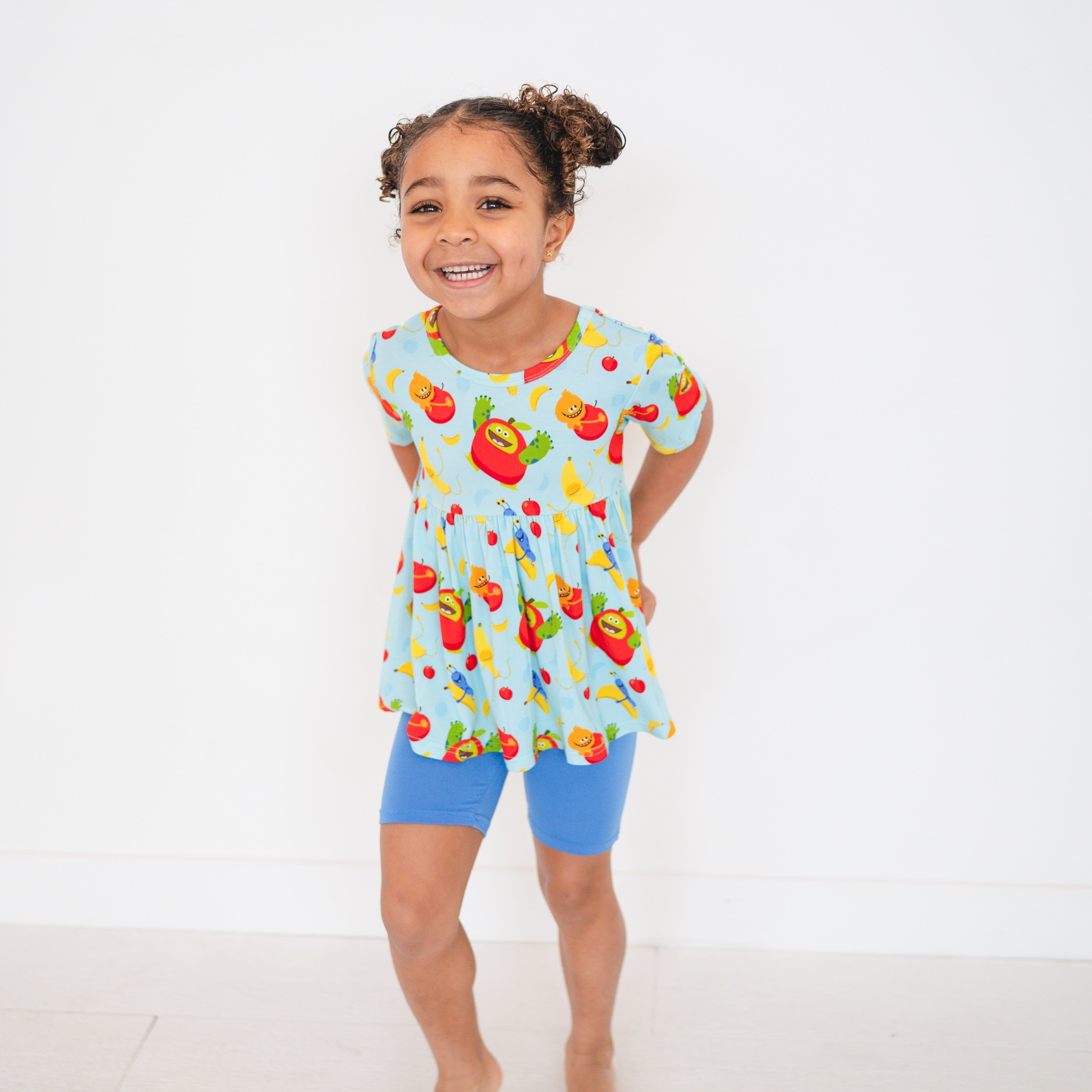 A young girl smiles in a light blue peplum top with a fruit monster print and blue biker shorts.