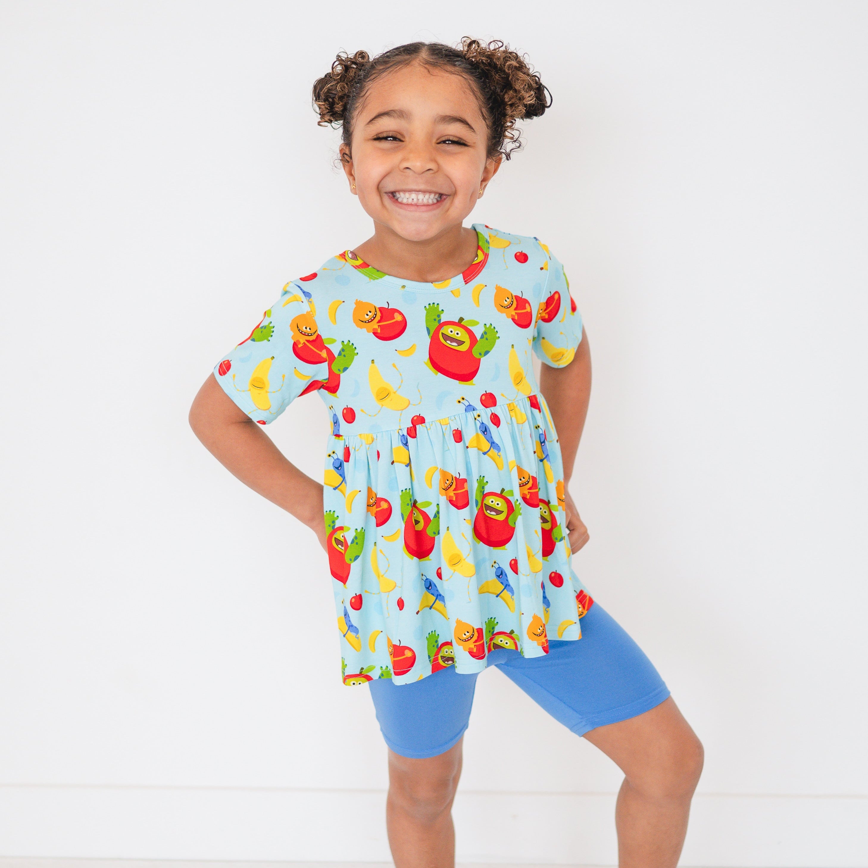 A young girl smiles in a light blue peplum top with a fruit monster print and blue shorts.