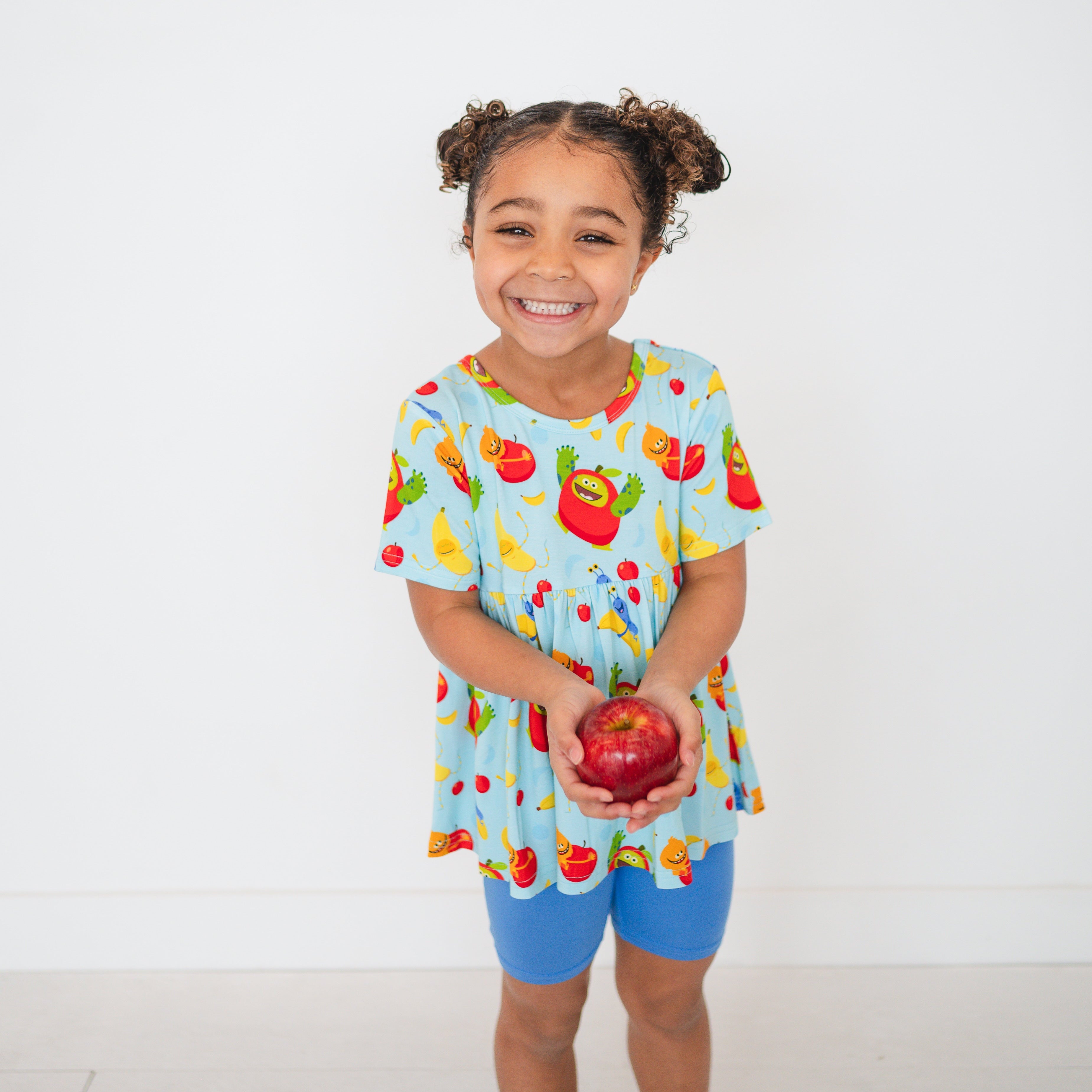 A young girl smiles holding a red apple, wearing a blue peplum top with fruit characters.