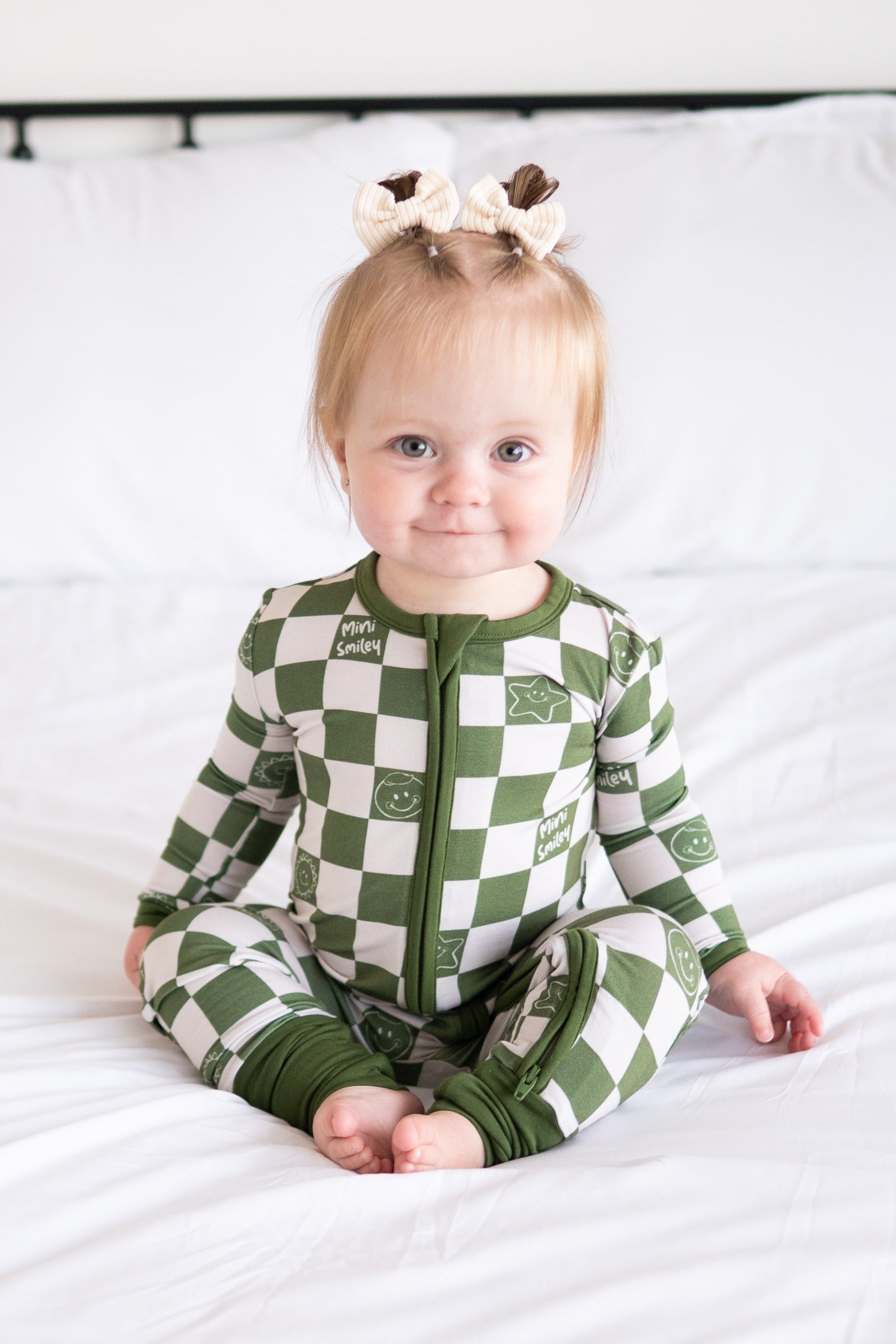 A baby girl sits on a white bed wearing a green and white checkered pajama set with smiley face prints.