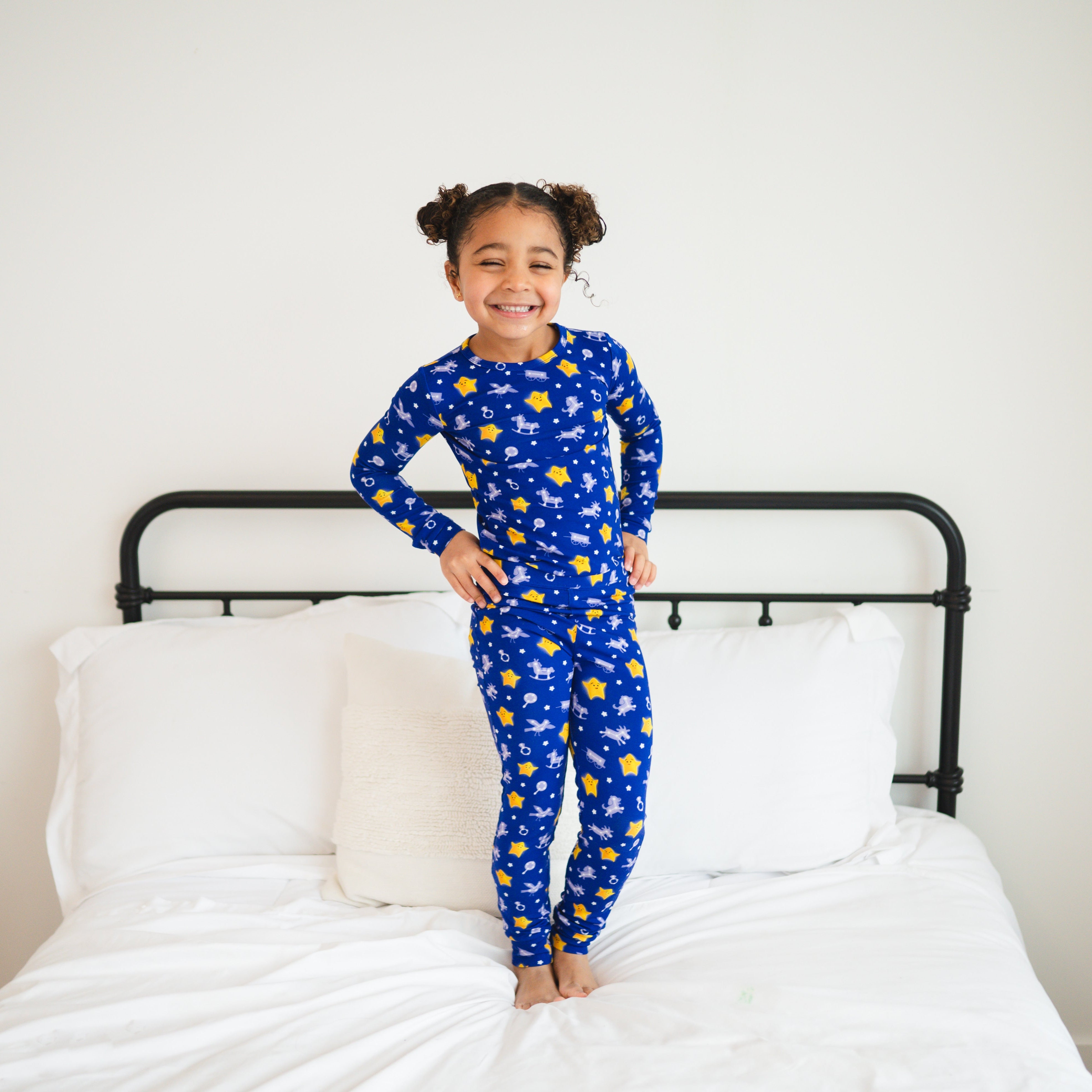 A young girl in blue pajamas with stars and toys smiles on a bed.