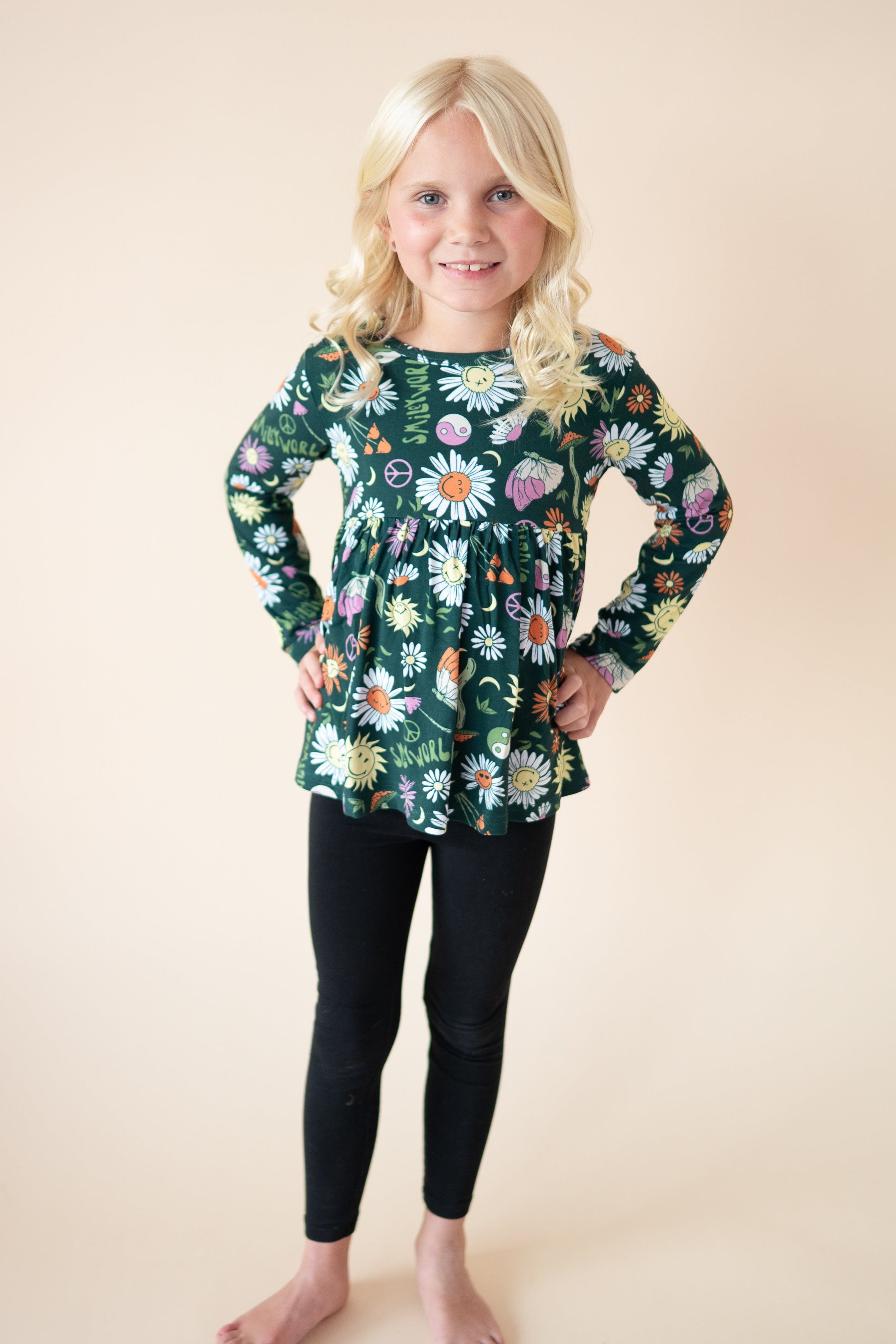 A young girl wears a long-sleeved green peplum top with a floral and smiley face print and black leggings.