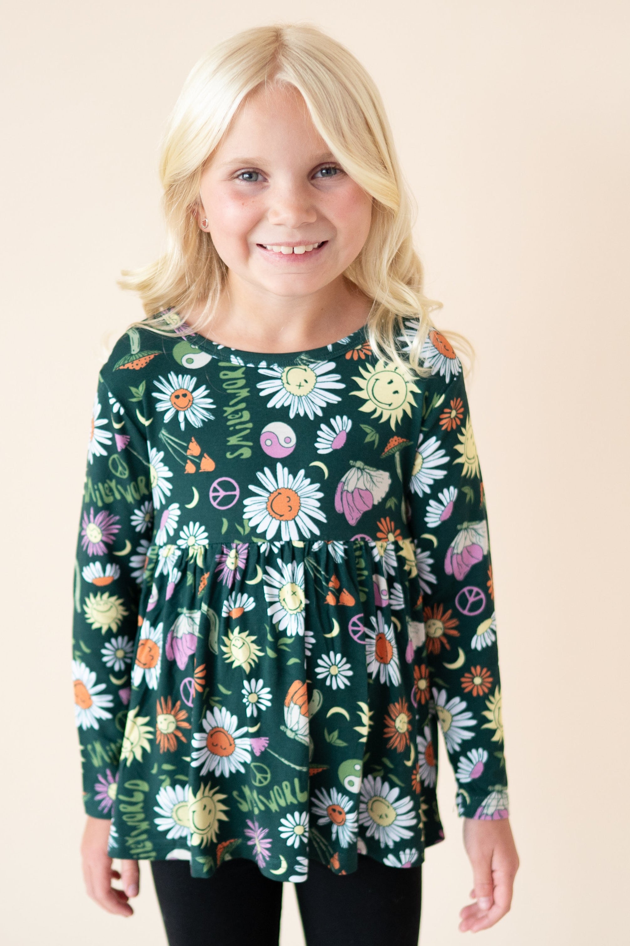 A young girl smiles wearing a long-sleeved green peplum top with a SmileyWorld floral print.