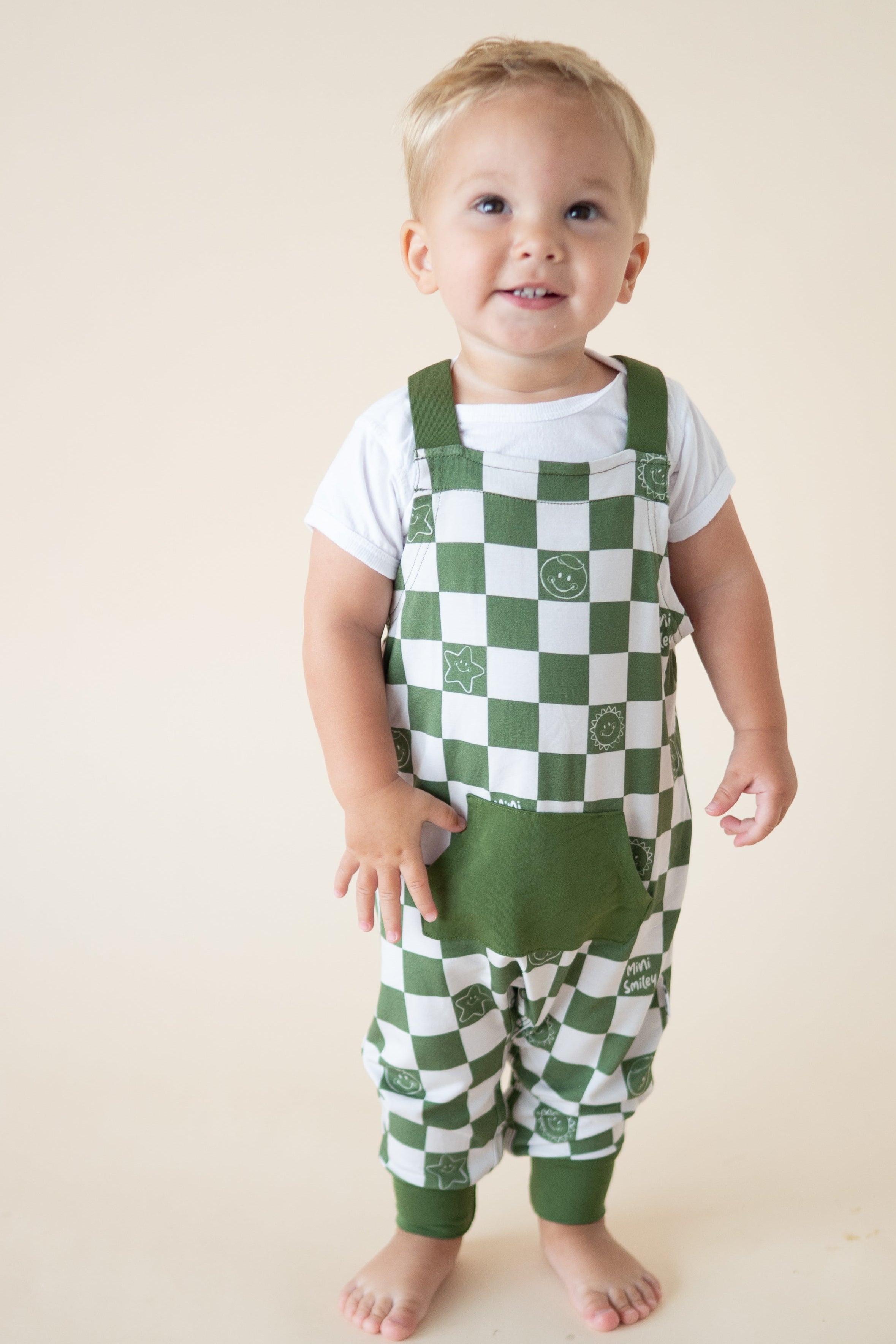 A baby boy wears green and white checkered overalls with smiley faces and stars over a white t-shirt.