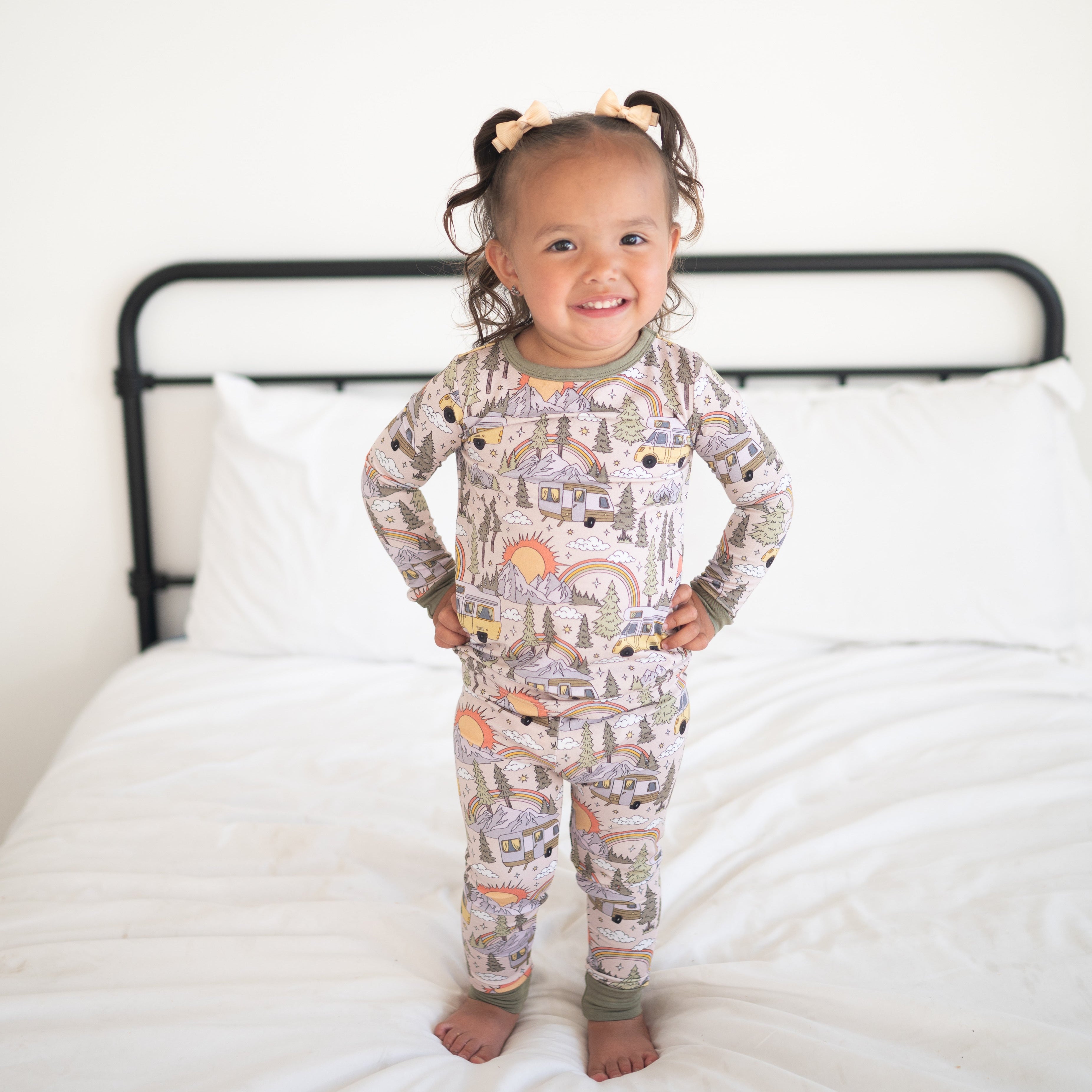 A young girl stands on a bed wearing a pink pajama set with a vintage camper print.