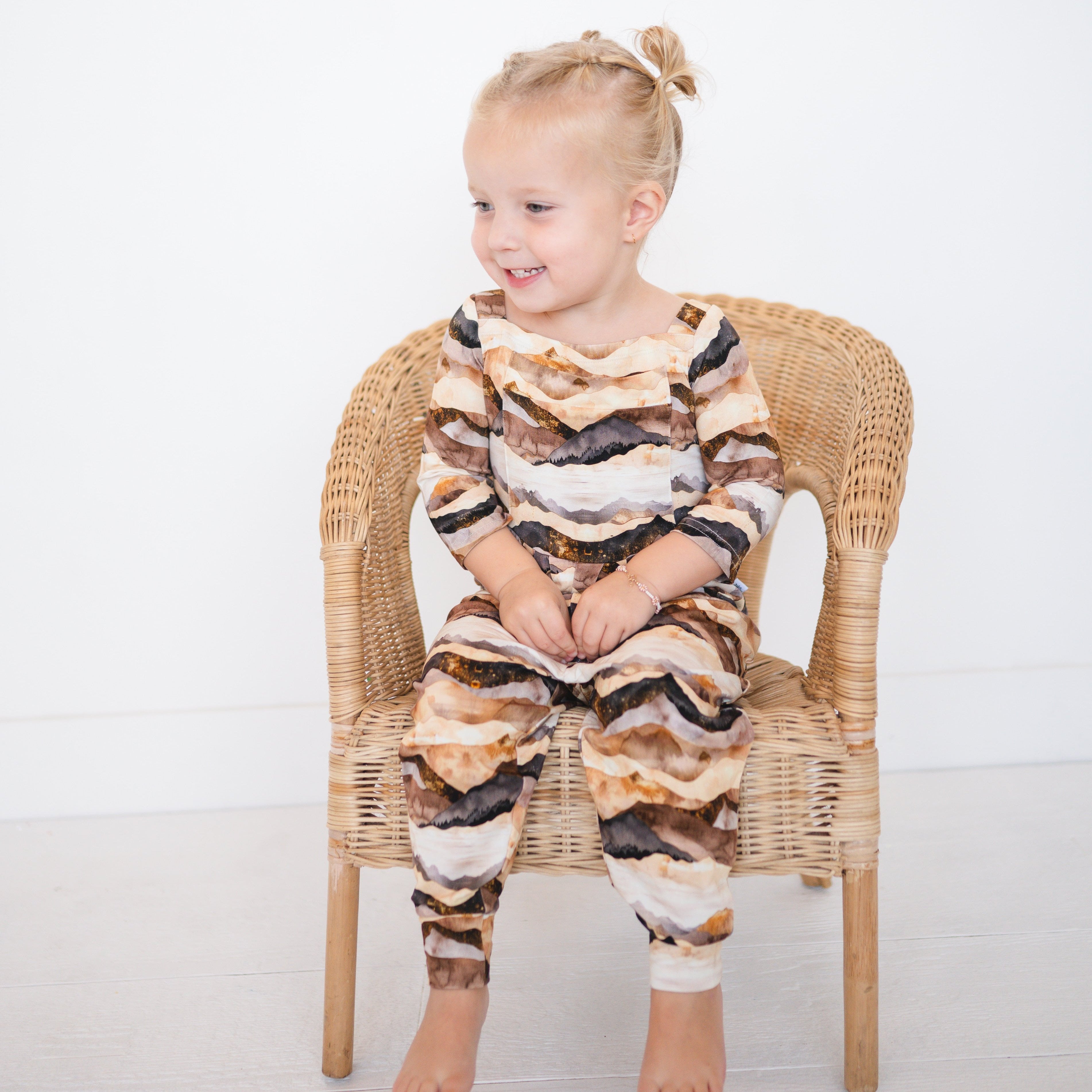 A young girl in a mountain range print bamboo romper sits on a wicker chair.