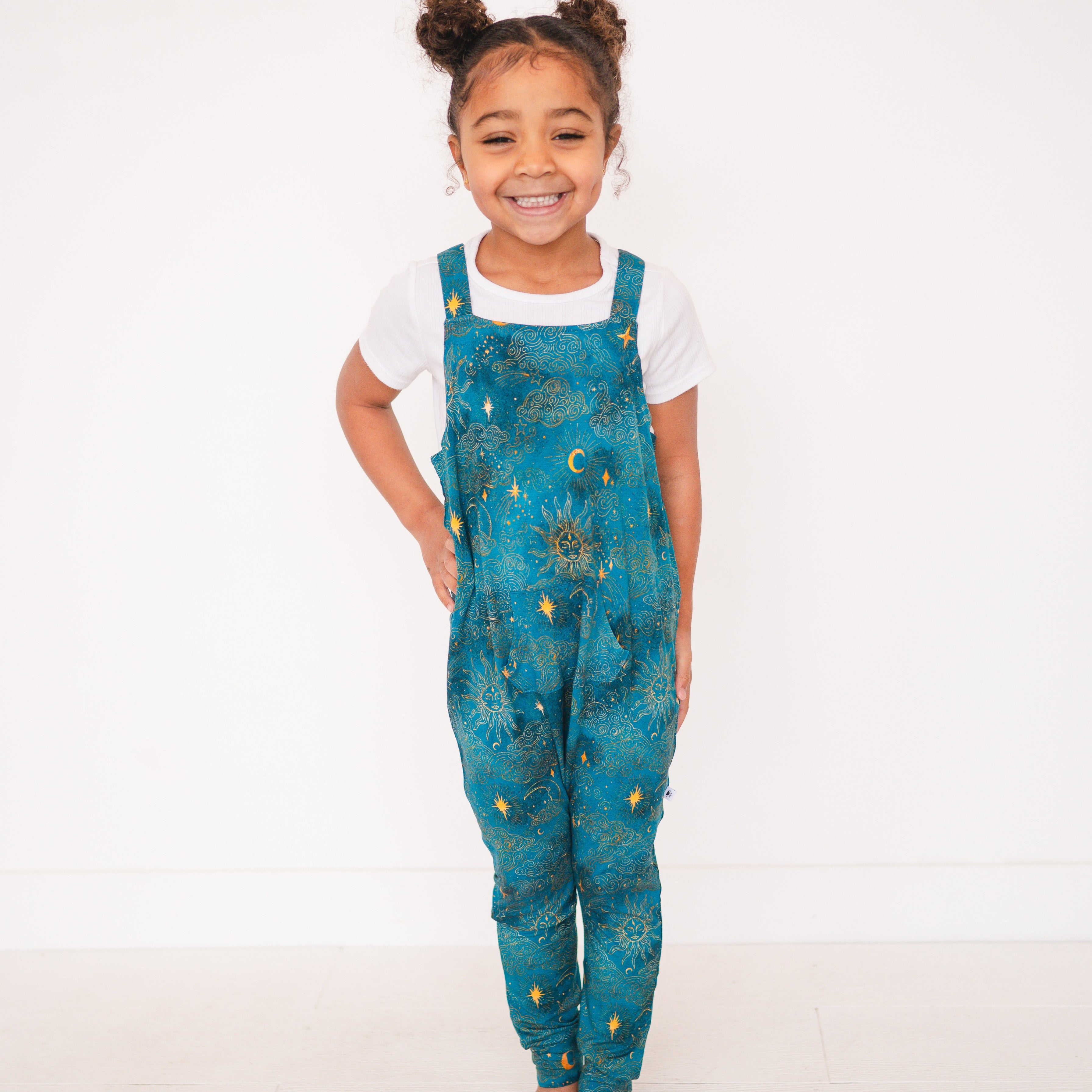 A young girl smiles wearing teal overalls with a celestial sun and moon print.