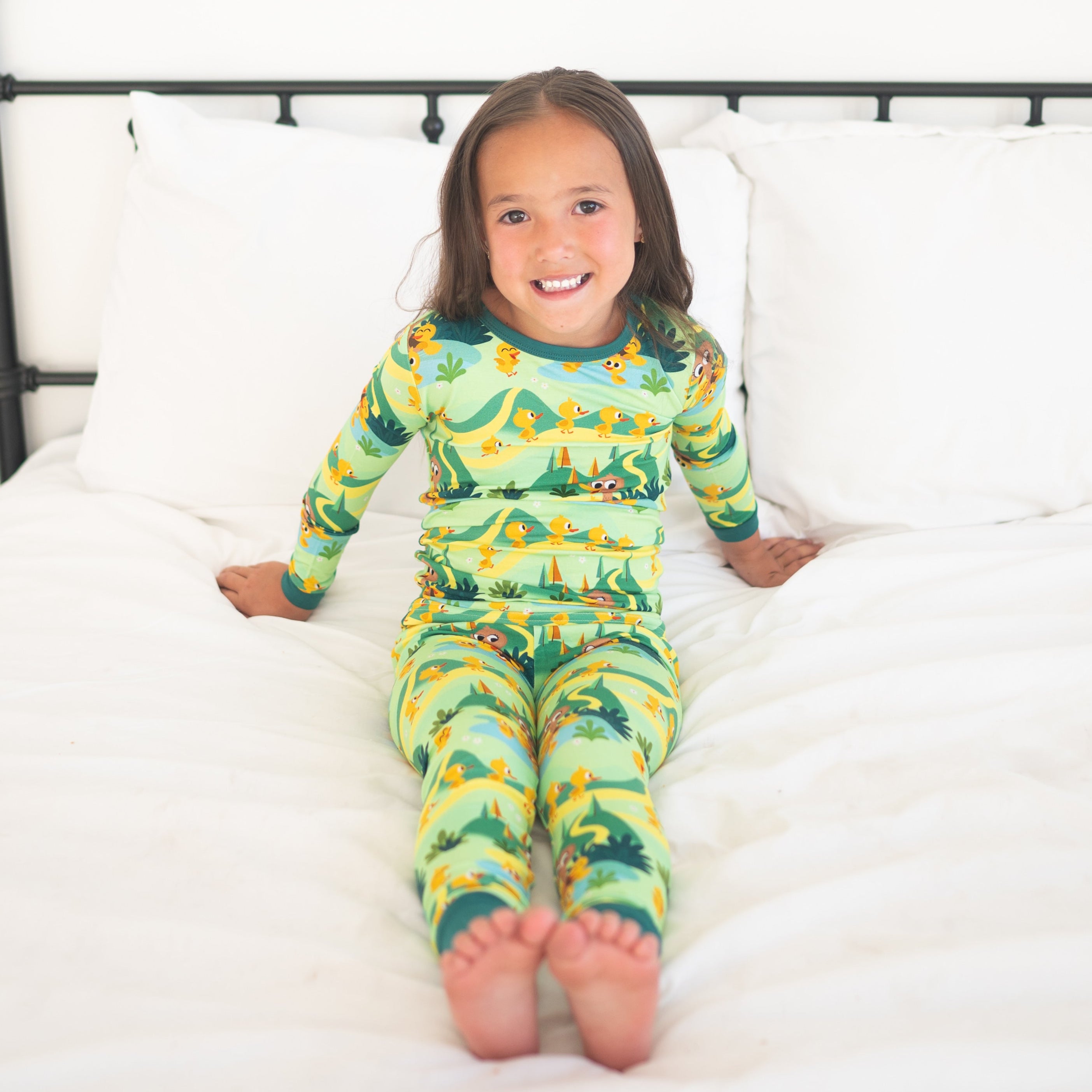 A young girl in green pajamas with a duck print sits on a white bed.
