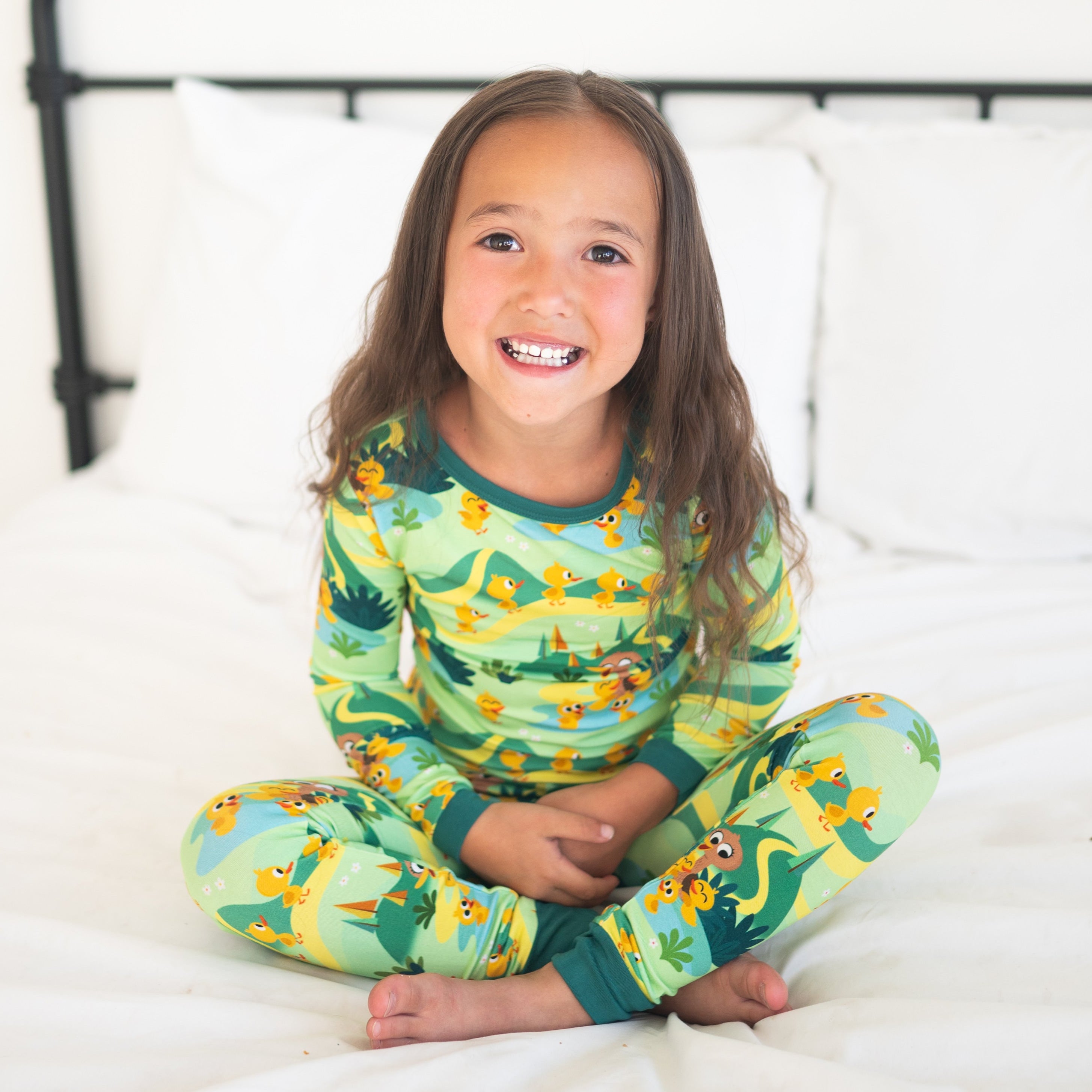 A young girl in green pajamas with a duck print sits on a bed.