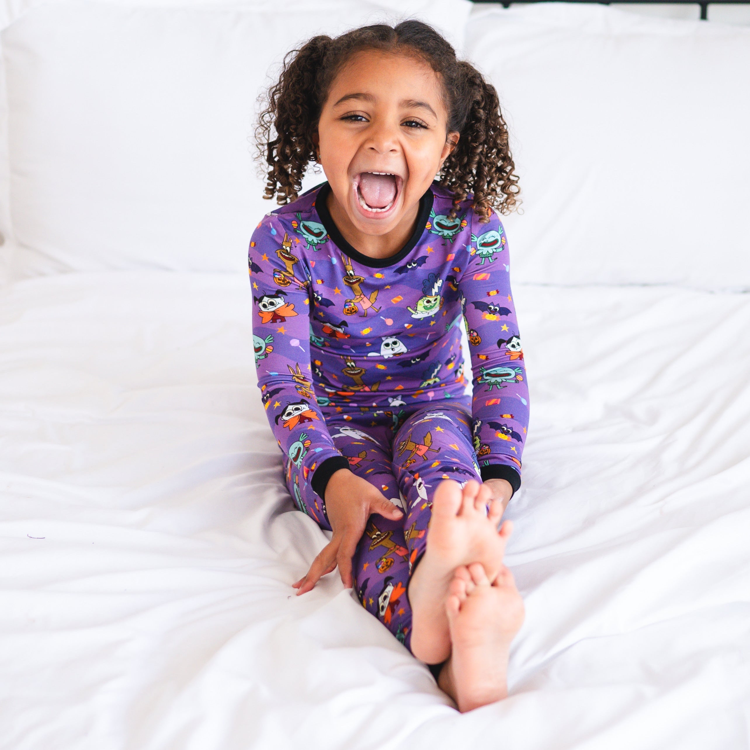 A young girl in purple monster-themed pajamas sits on a white bed.