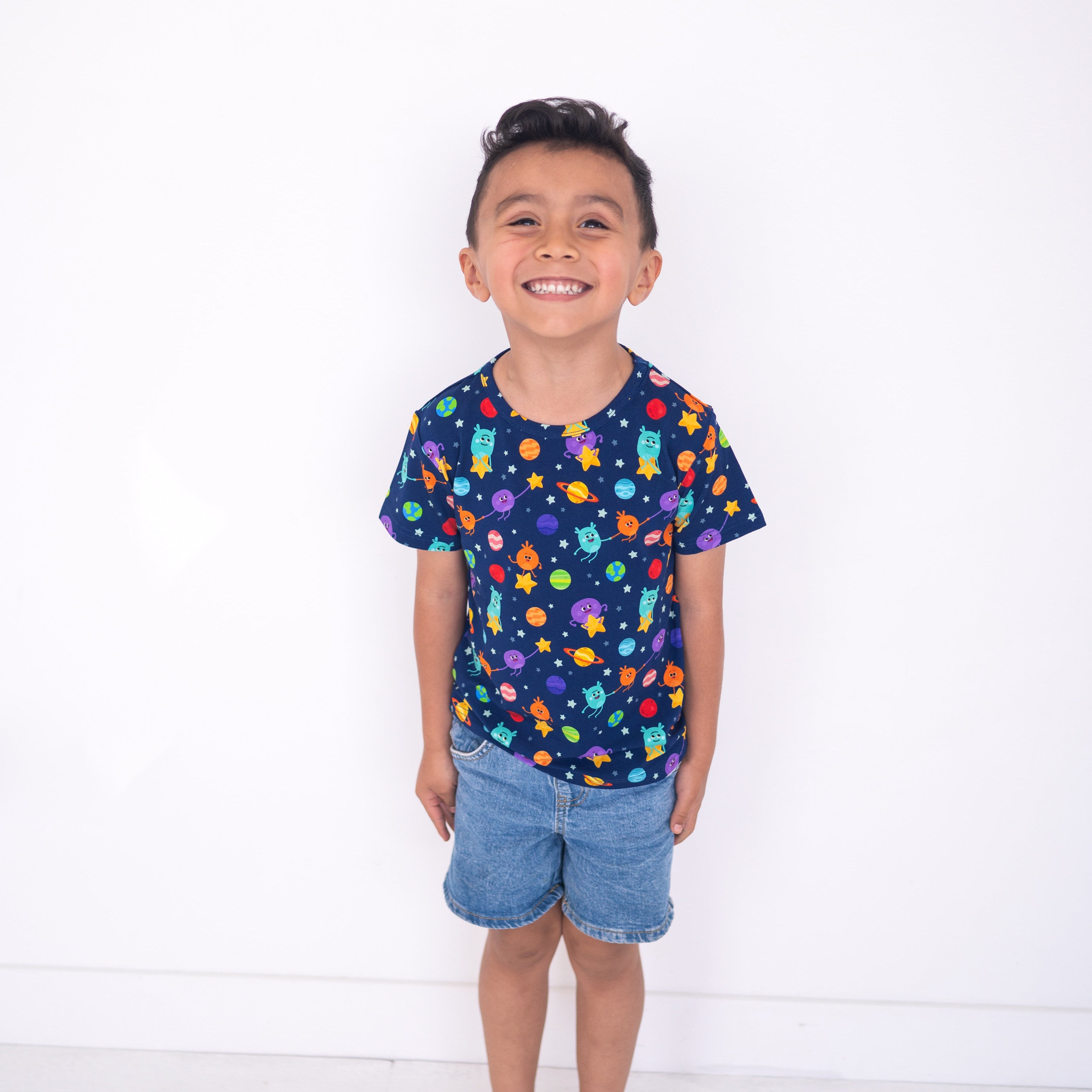 A young boy smiles wearing a navy blue t-shirt with a colorful space-themed print and denim shorts.