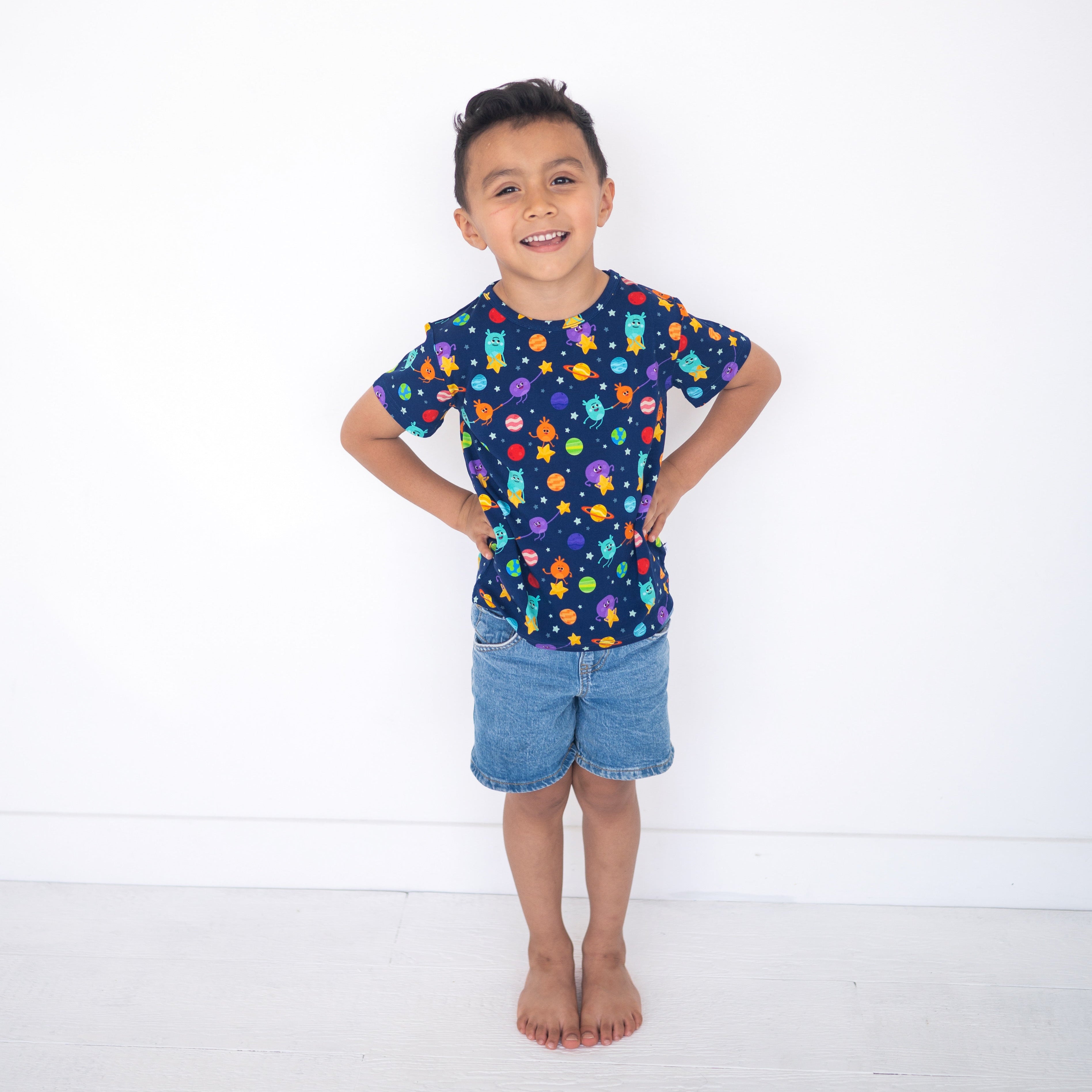 A young boy smiles while wearing a navy blue t-shirt with a space-themed print and denim shorts.