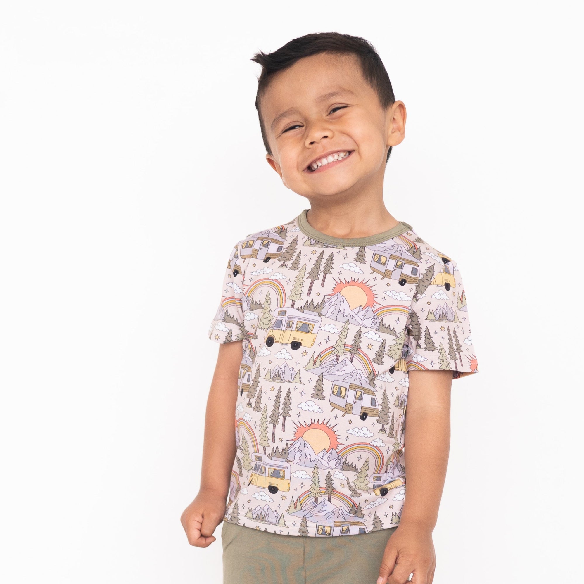 A young boy smiles wearing a light pink t-shirt with a vintage camper and mountain print.