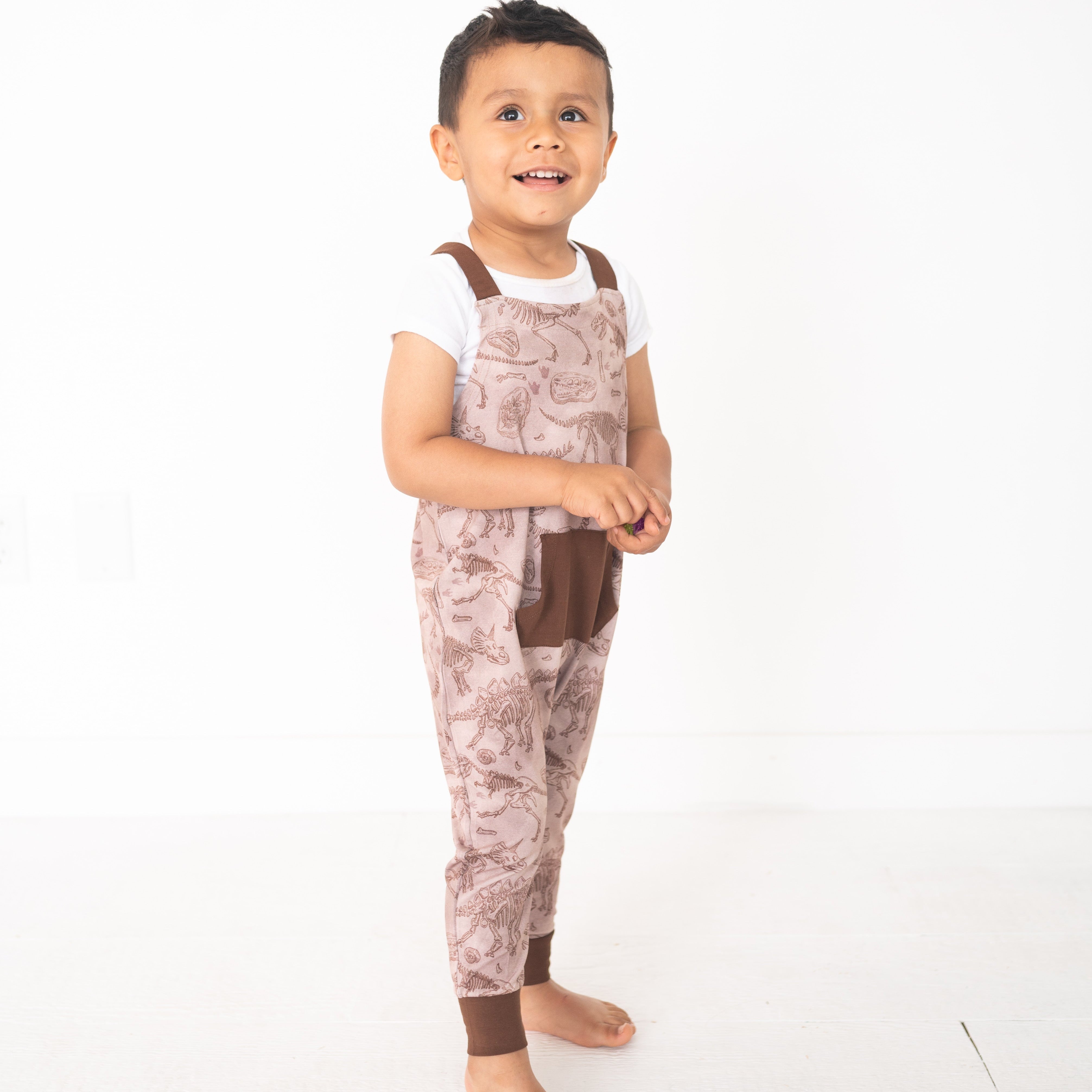A young boy wears brown fossil-print overalls over a white t-shirt.