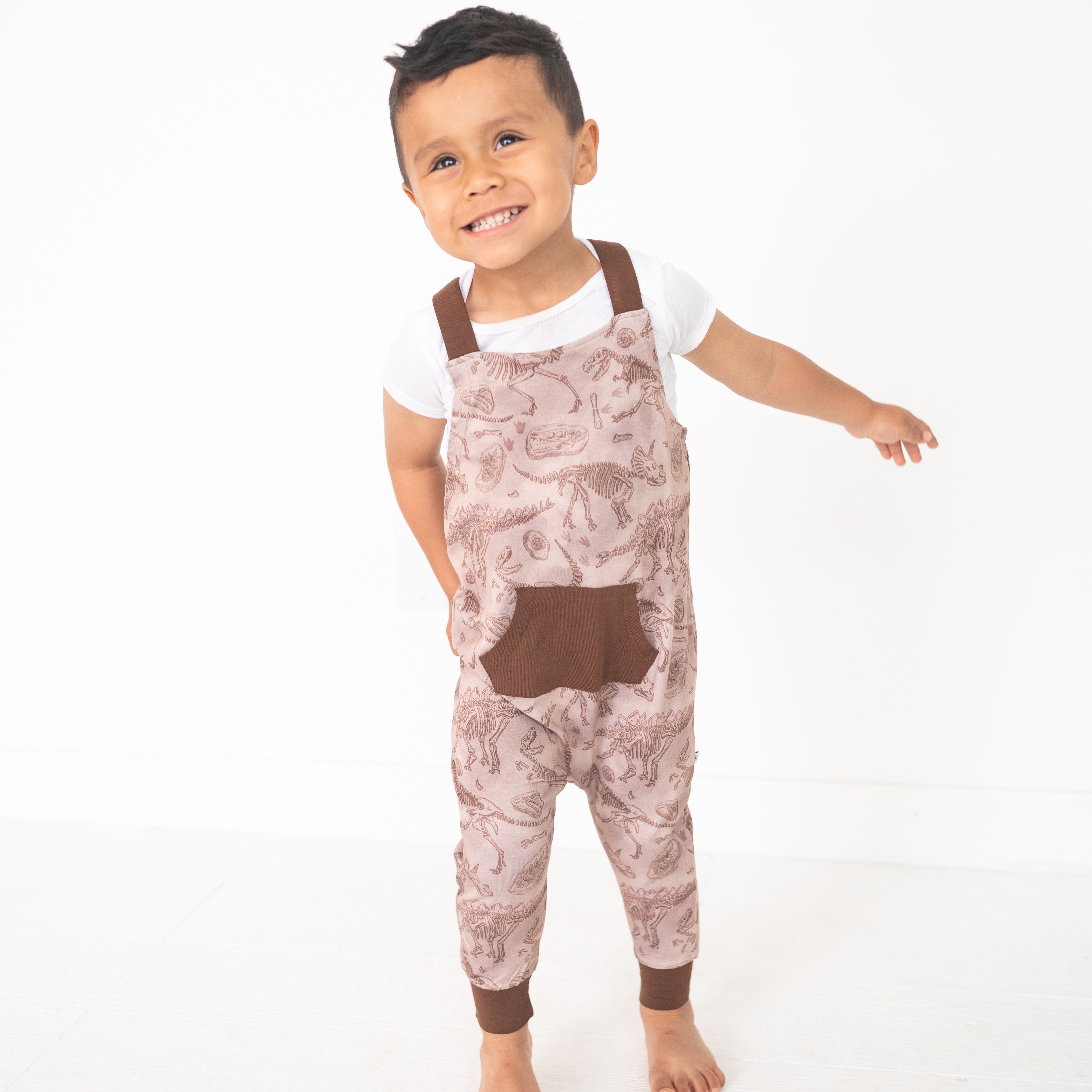 A young boy smiles wearing brown fossil-print overalls over a white t-shirt.