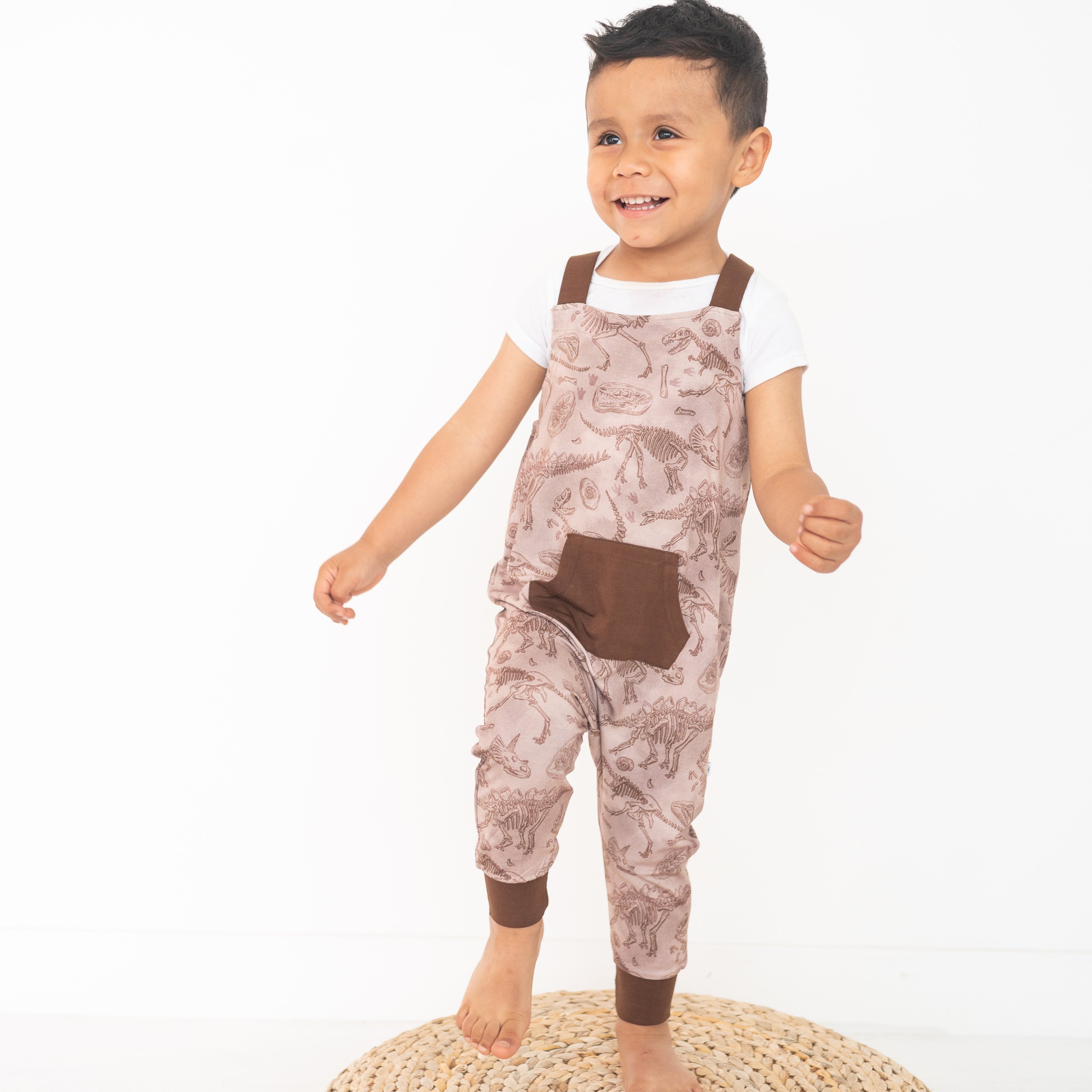 A young boy wears brown dinosaur-print overalls over a white t-shirt.