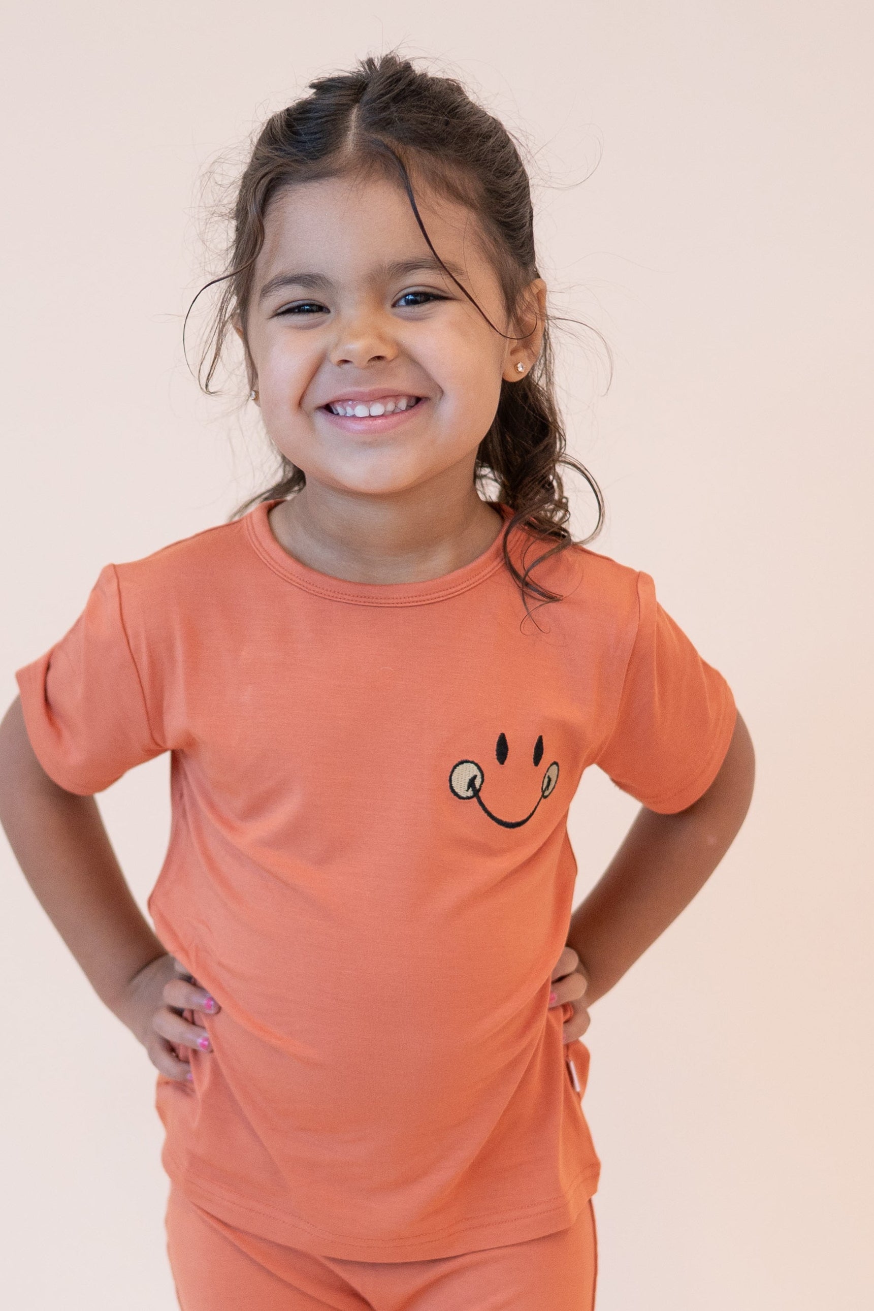 A young girl smiles wearing an orange t-shirt with a smiley face graphic.
